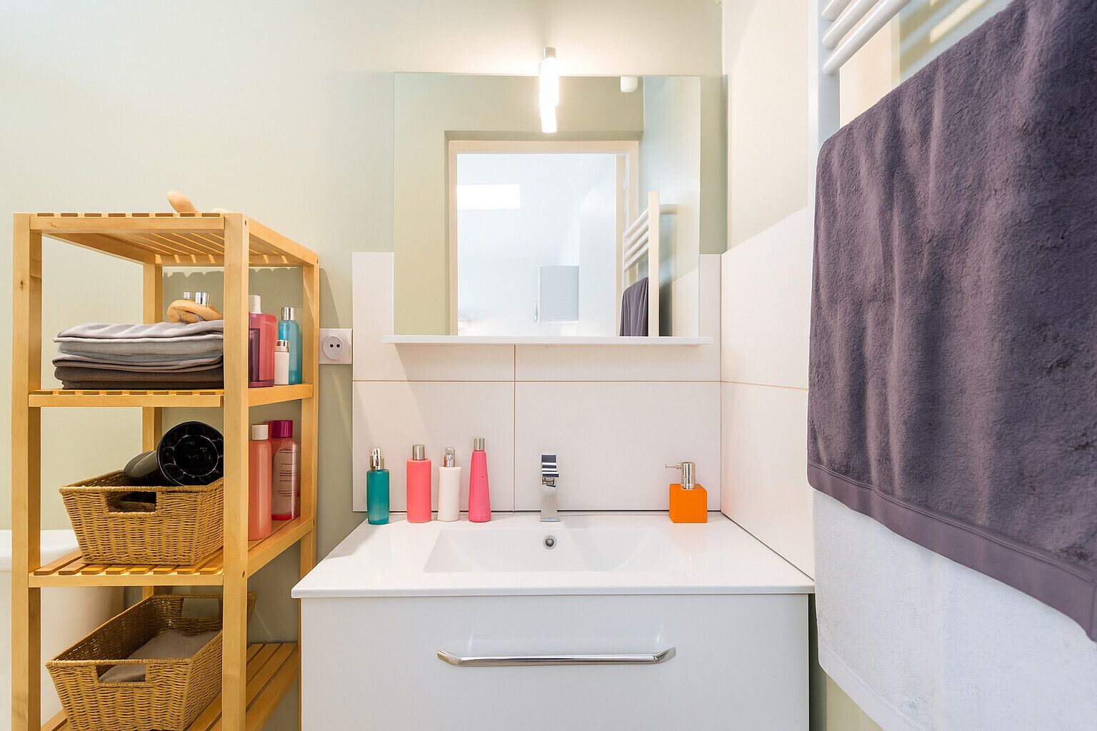 Un baño con estante de madera que contiene toallas y artículos de aseo, un lavamanos blanco con espejo encima, y una toalla morada colgada en la pared.