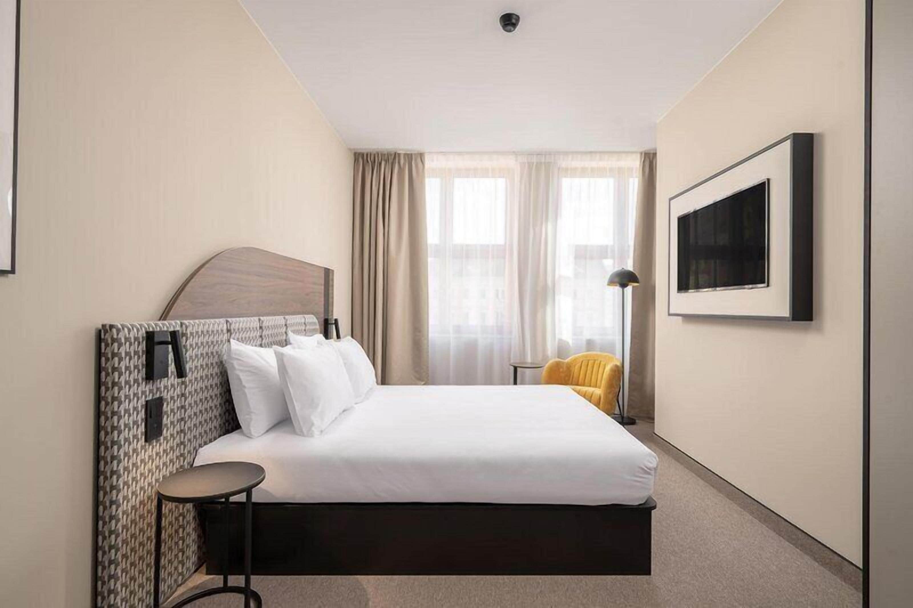A hotel room with a bed, a chair, a desk, a lamp, and a window with curtains.