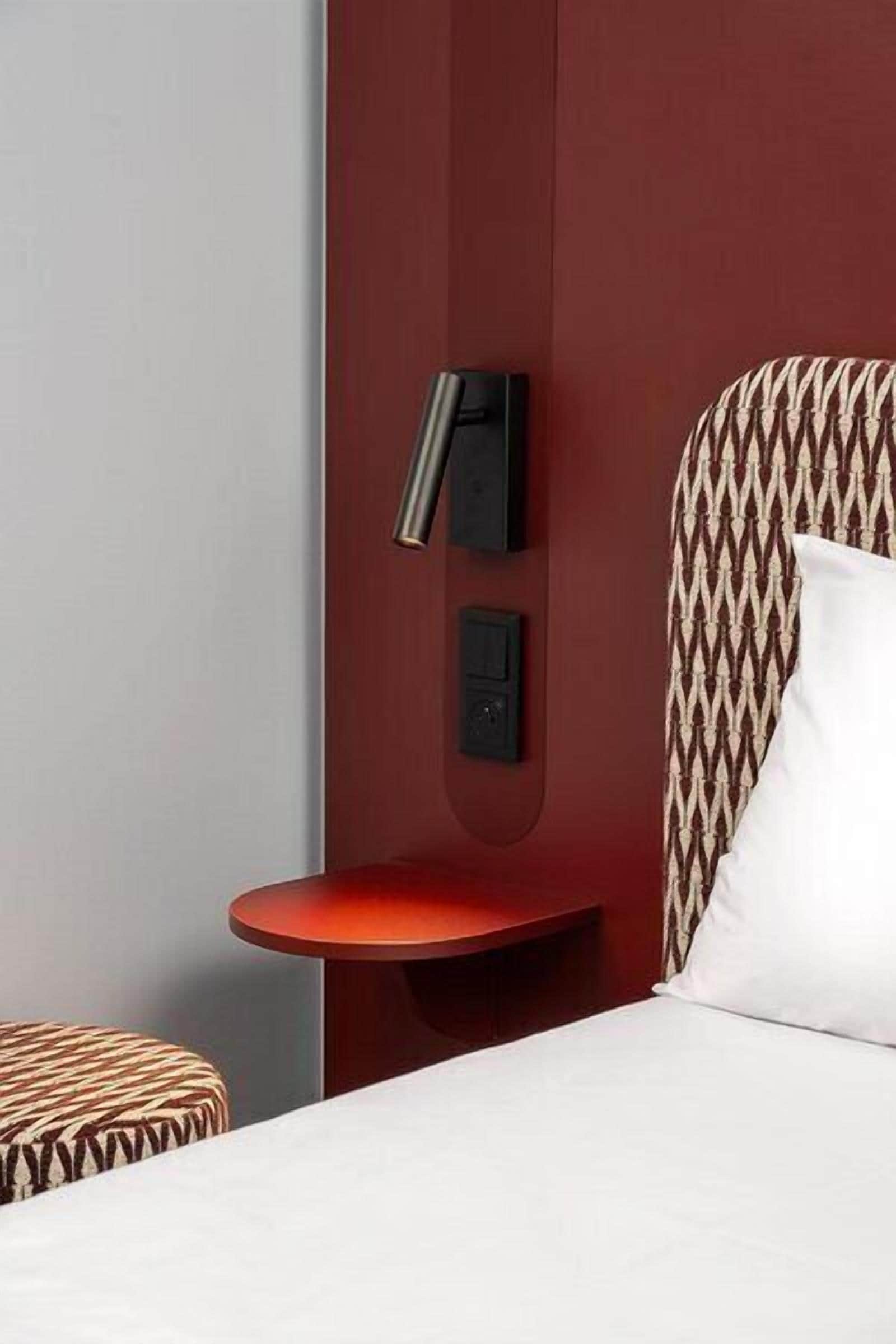 A hotel room with a bed, a red side table, a wall-mounted control panel, and a patterned headboard.