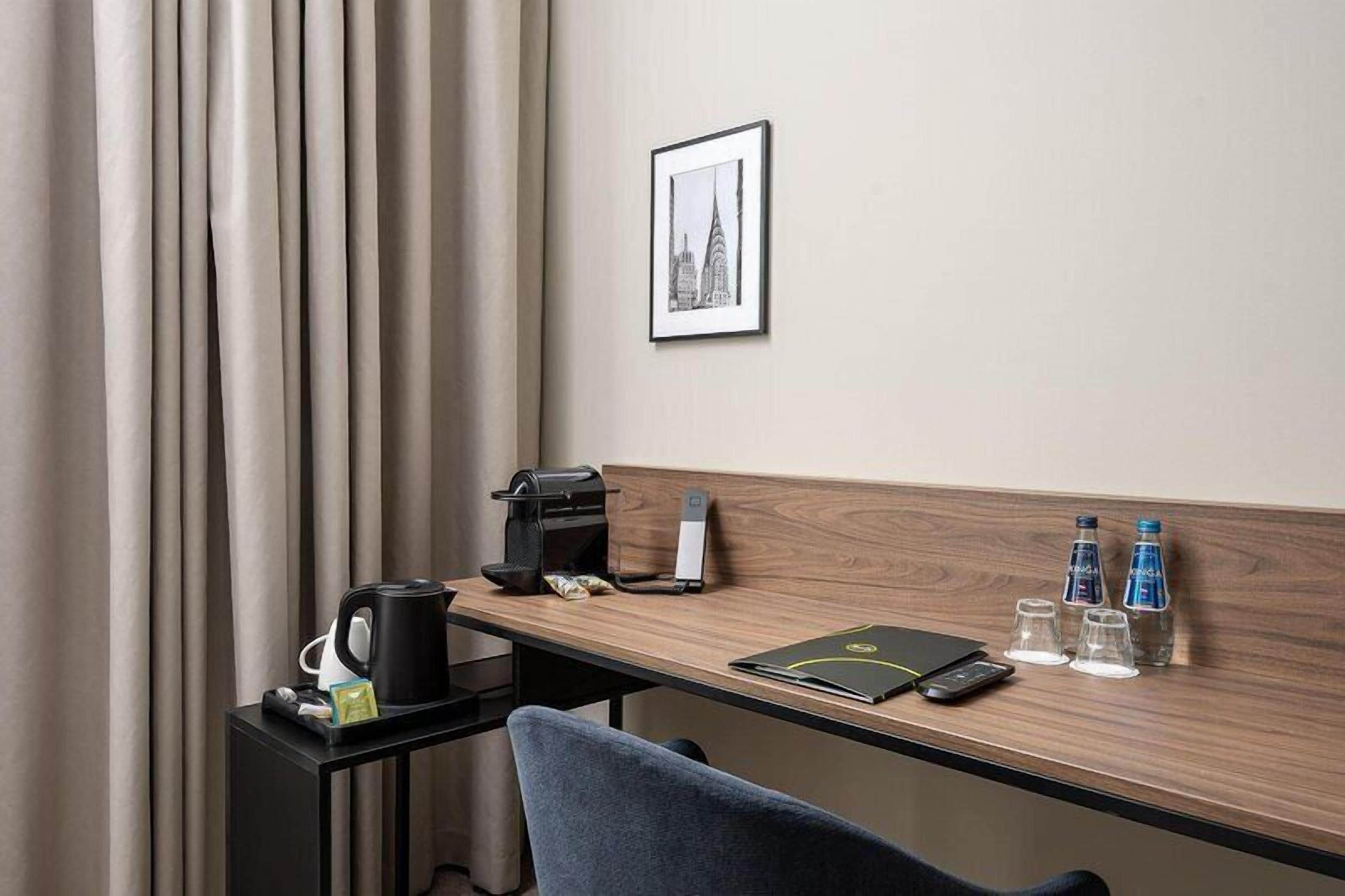 Habitación de hotel con un escritorio de madera, una cafetera negra, un teléfono inteligente, una libreta y dos botellas de agua.
