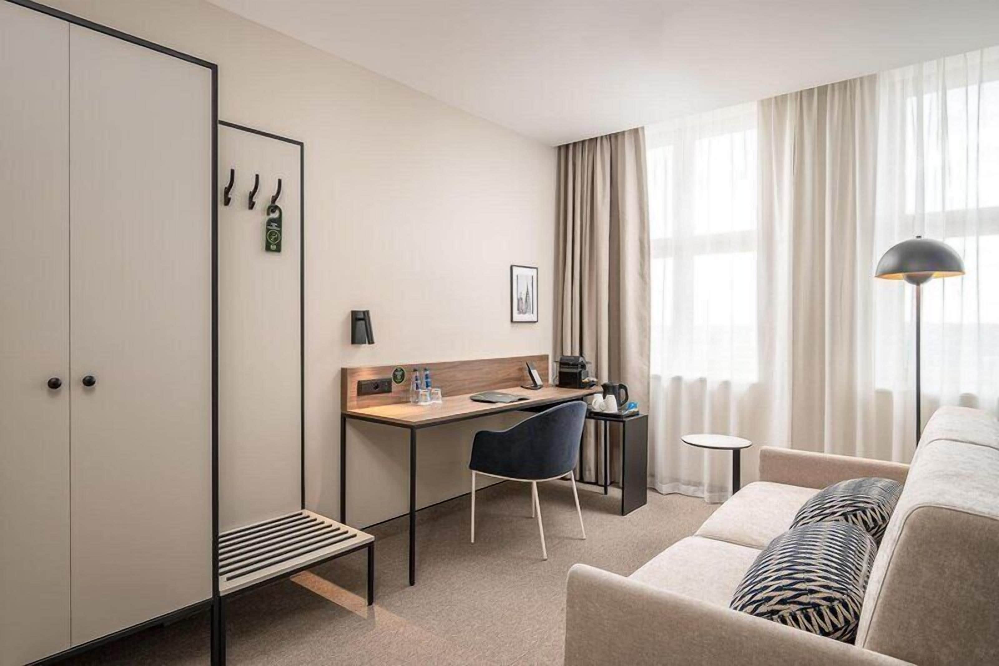 A hotel room with a bed, a desk with a chair, a small table, a lamp, and a wardrobe.