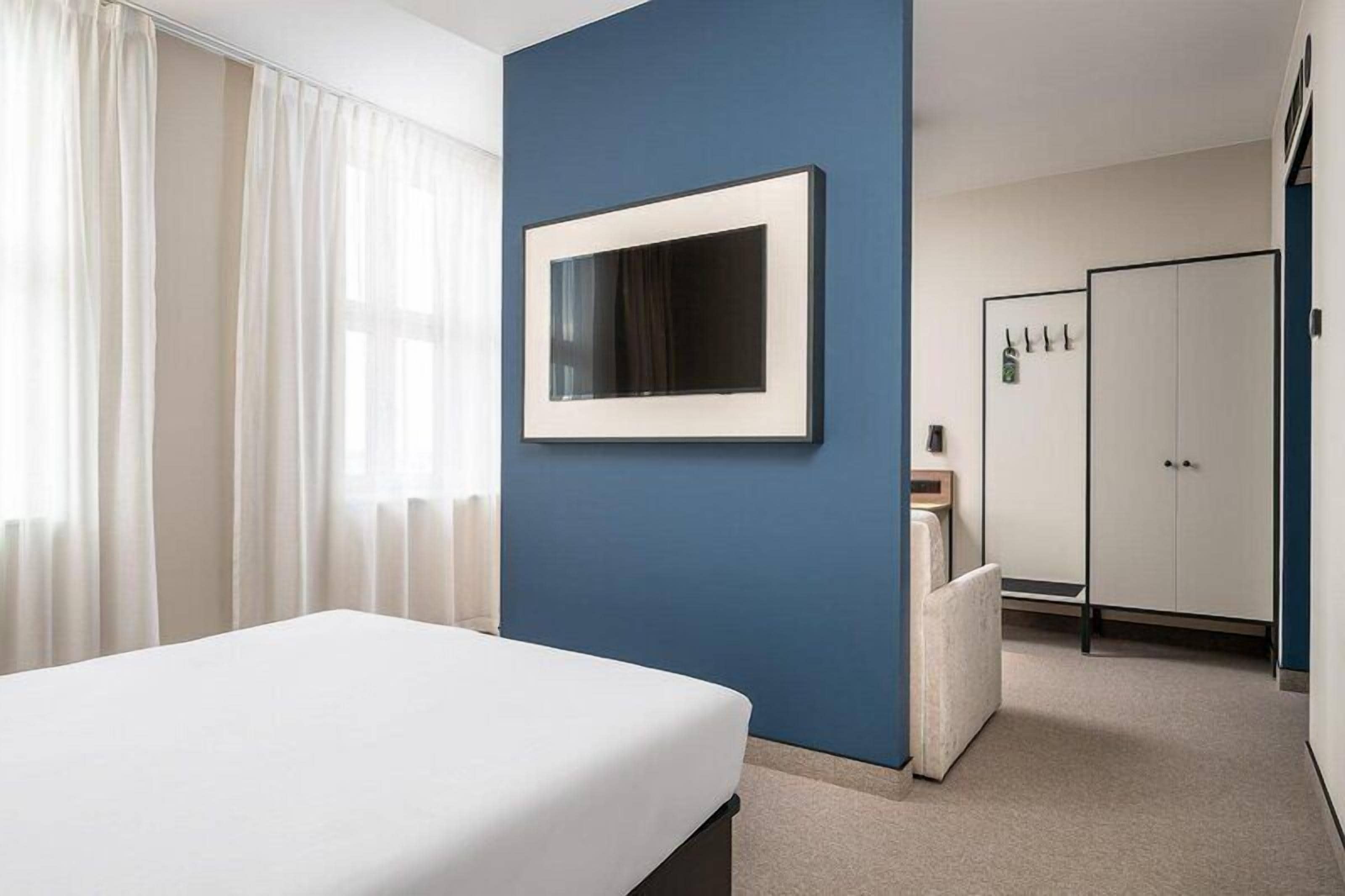 Une chambre d’hôtel avec un lit, un mur d’accent bleu, un miroir encadré, une armoire et un petit coin salon.