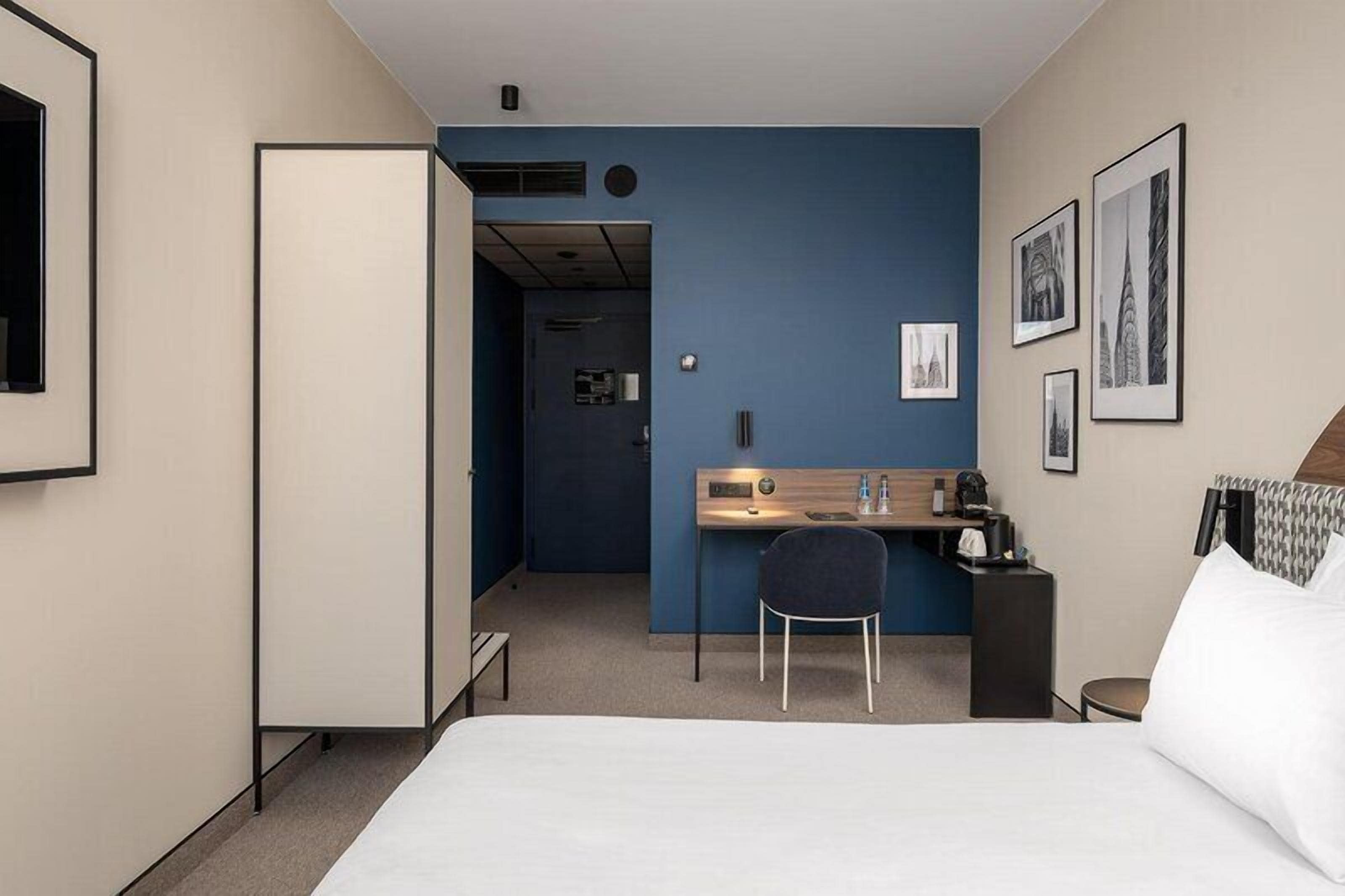 A hotel room with a bed, a desk, a chair, a wardrobe, and a door leading to another room.