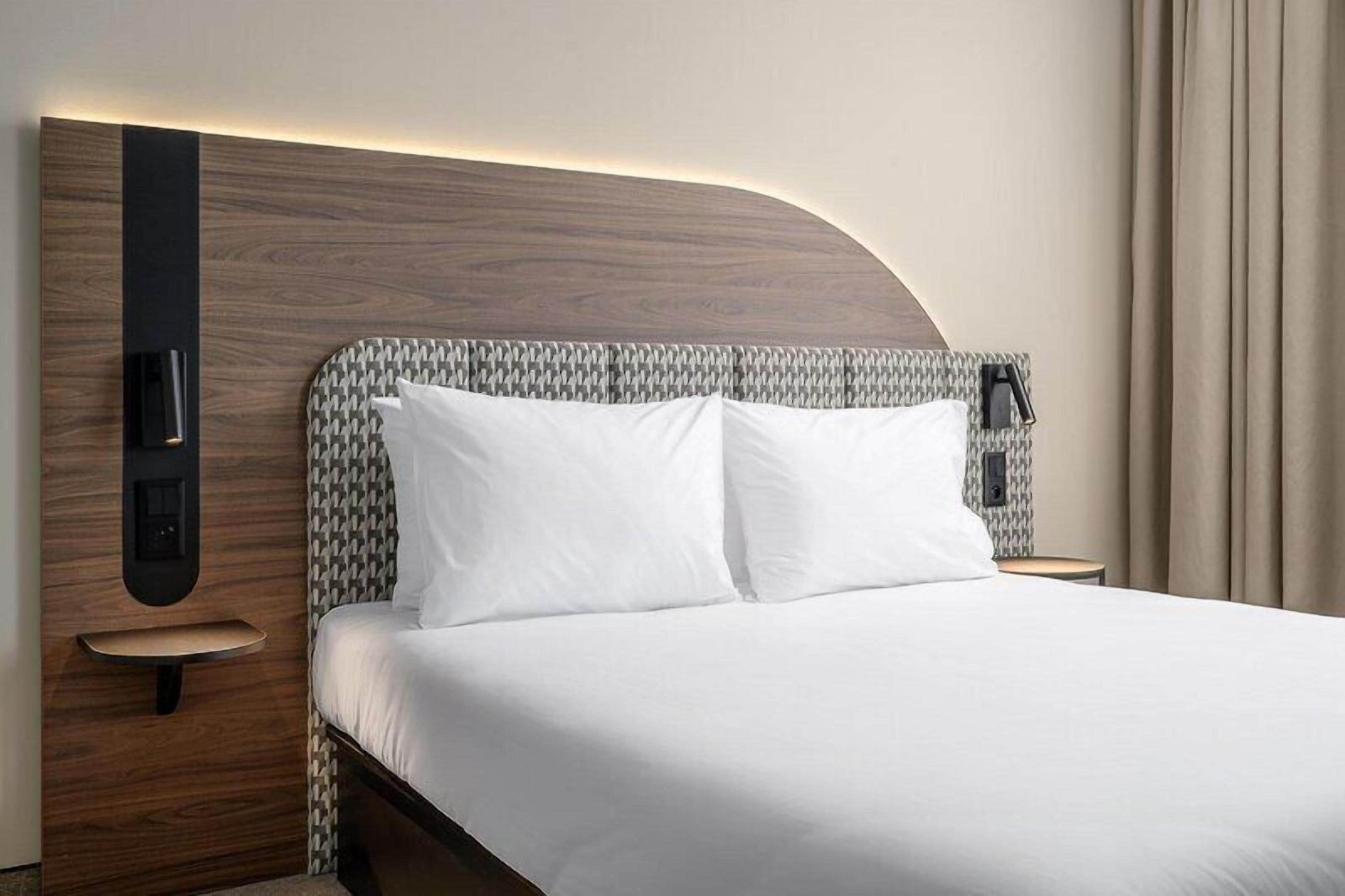 Una habitación de hotel con cama, mesita de noche, lámpara de pared y cortina.