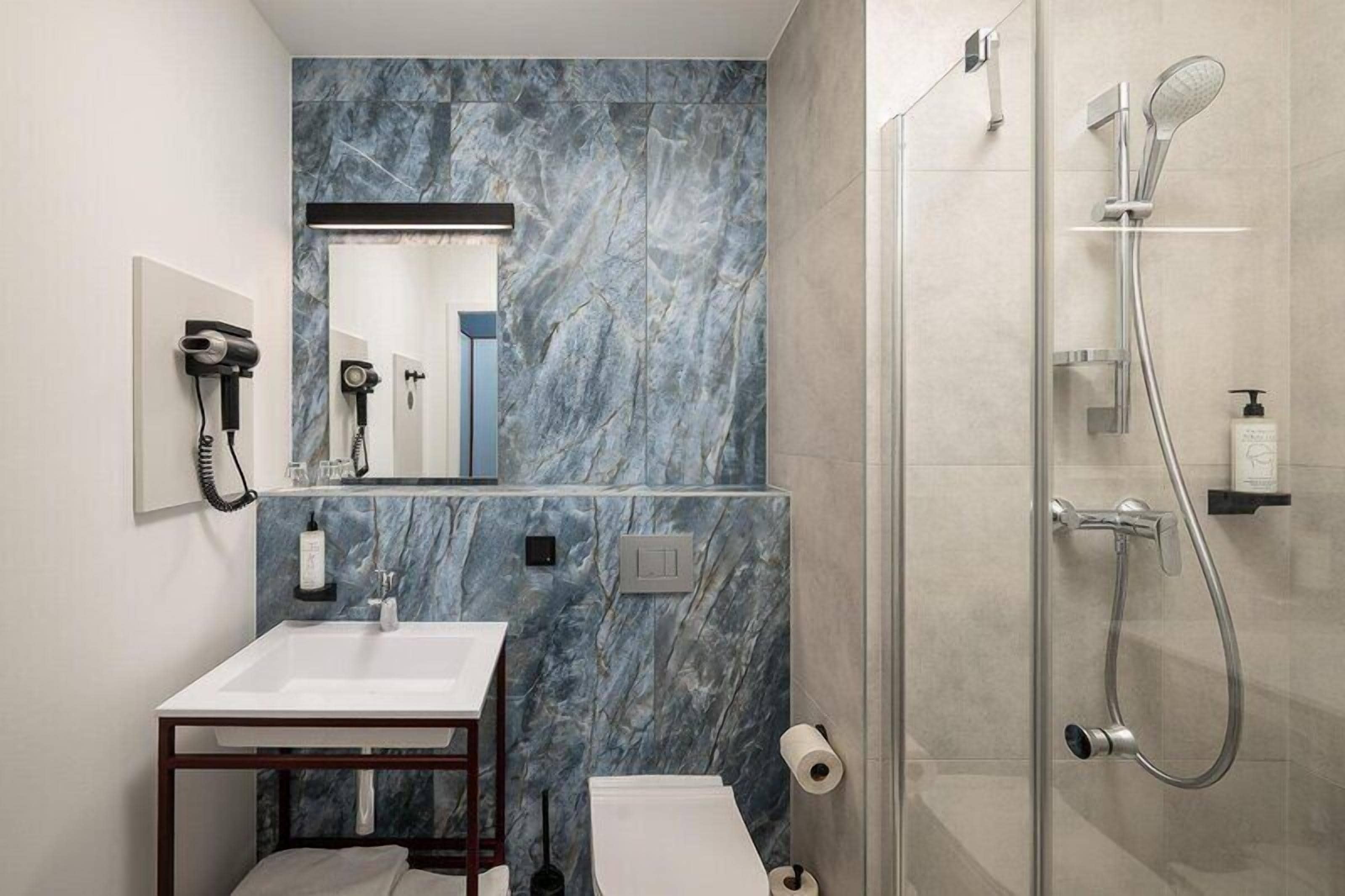 A bathroom with a marble wall, a sink, a mirror, a hairdryer, a shower, and a toilet.