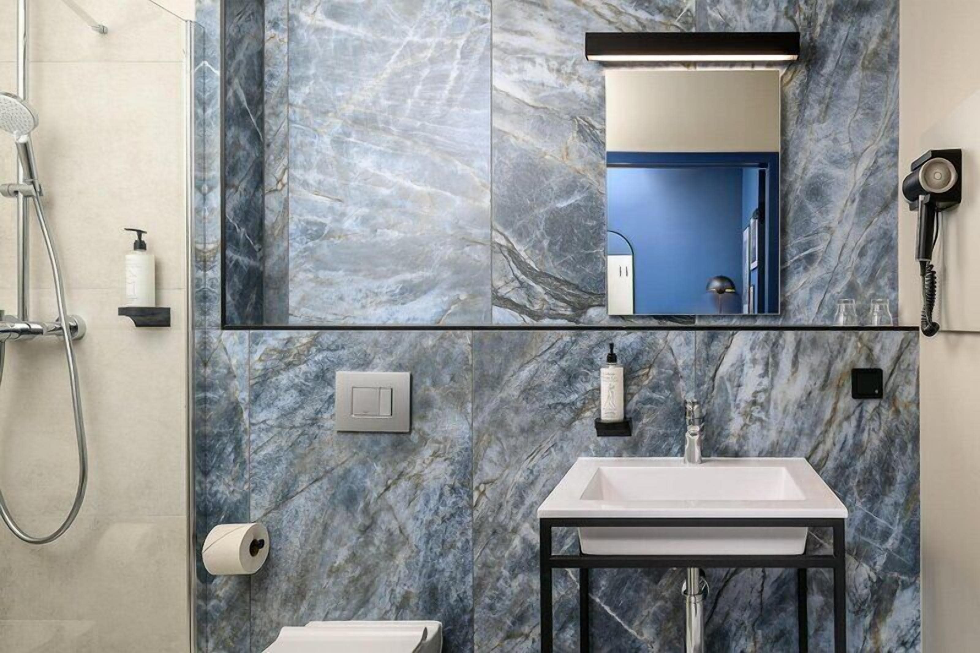 A bathroom with a marble wall, a sink, a mirror, a shower, and a toilet.