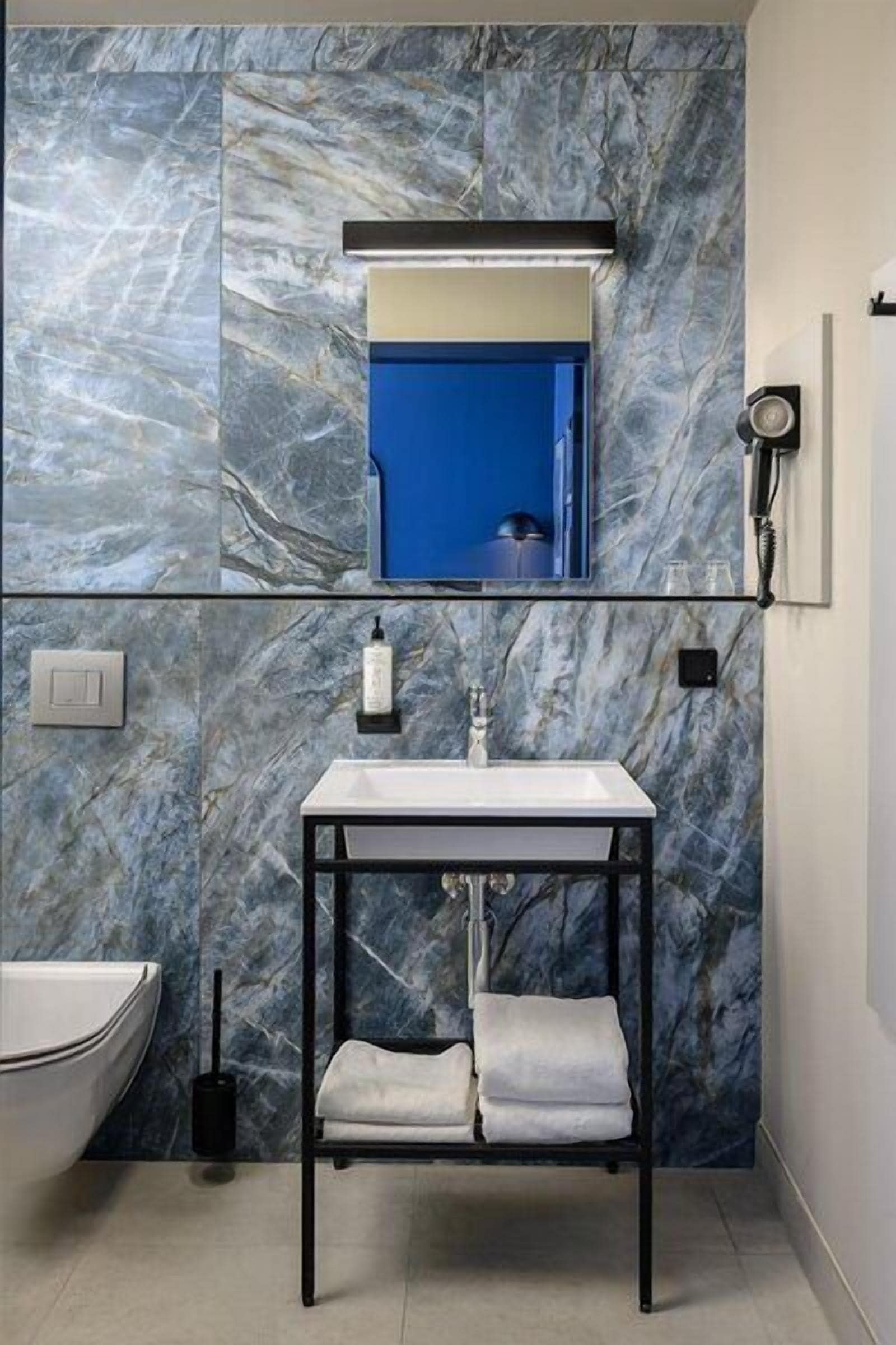 A bathroom with a marble wall, a sink, a mirror, a towel rack, and a handheld showerhead.
