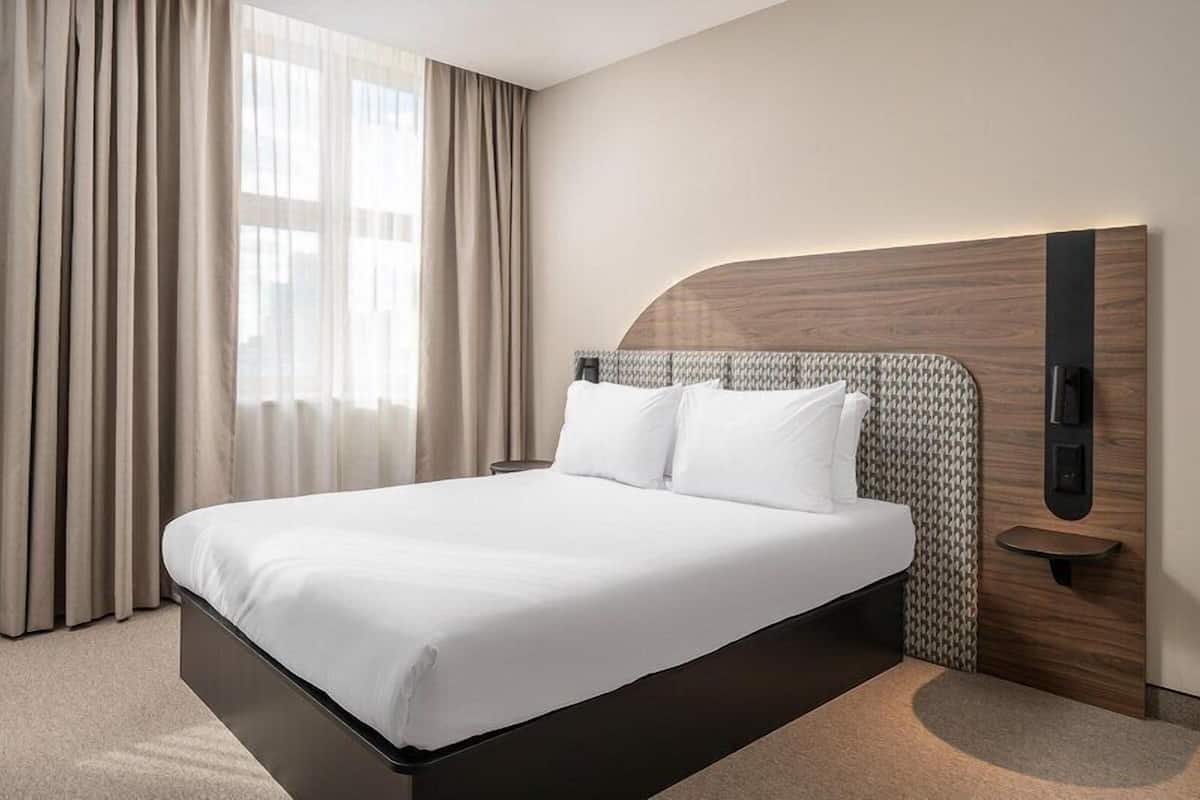 A hotel room with a large bed, a wooden headboard, a bedside table, and a window with curtains.