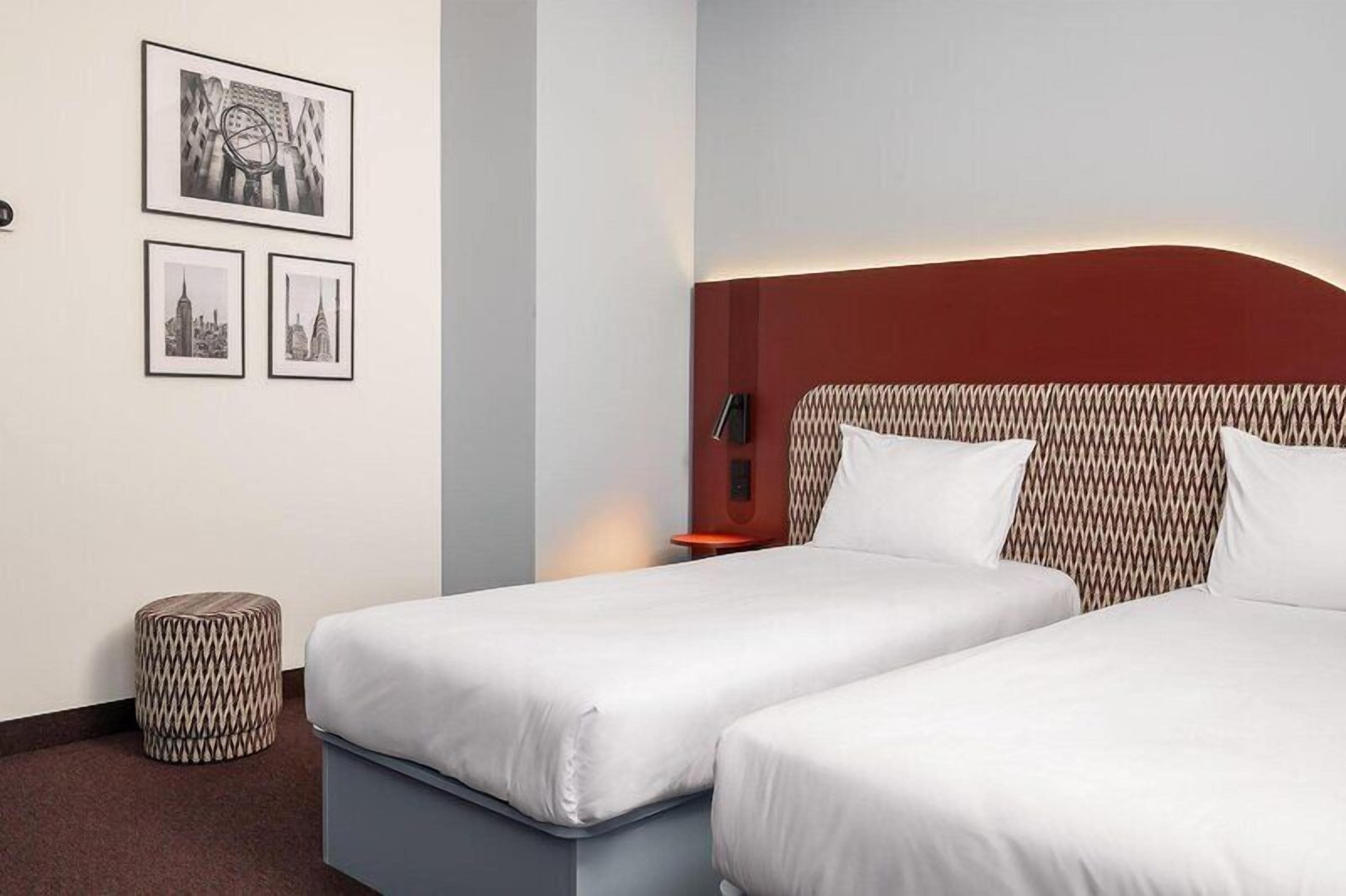 Habitación de hotel con dos camas individuales, cabecera estampada, cesto de basura y cuadros con vistas urbanas en la pared.