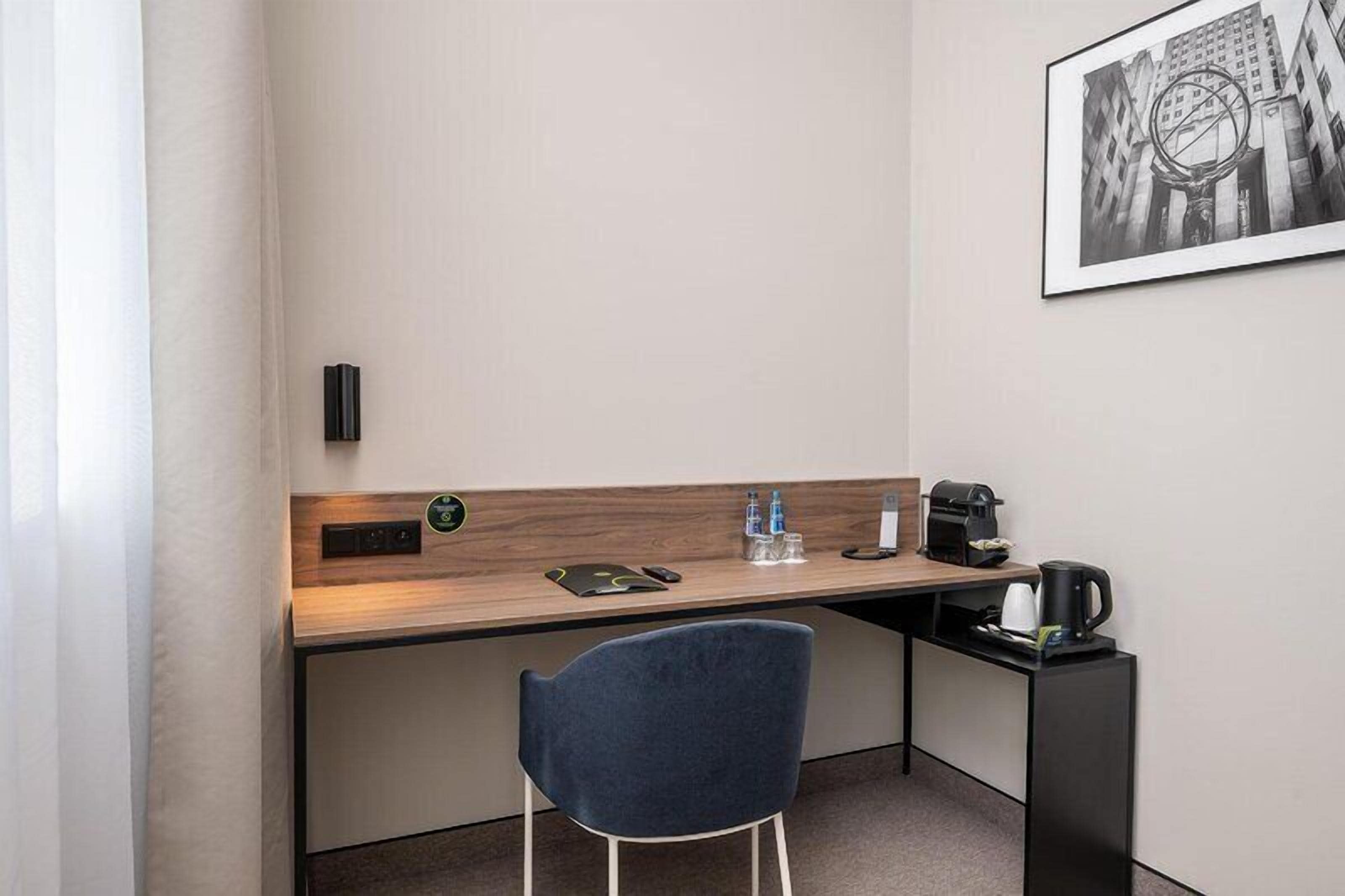 A hotel room with a desk, chair, coffee maker, water bottles, and a framed picture on the wall.