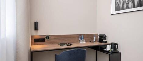 A hotel room with a desk, chair, coffee maker, water bottles, and a framed picture on the wall.