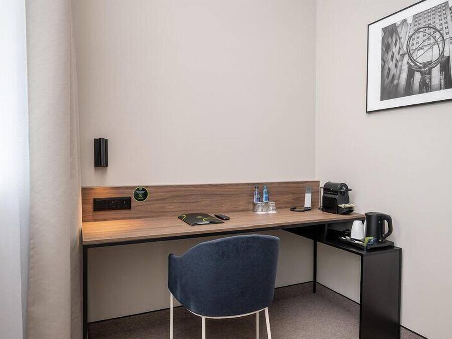 Hotellihuone, jossa on työpöytä, tuoli, kahvinkeitin, vesipulloja ja seinällä kehystetty kuva.