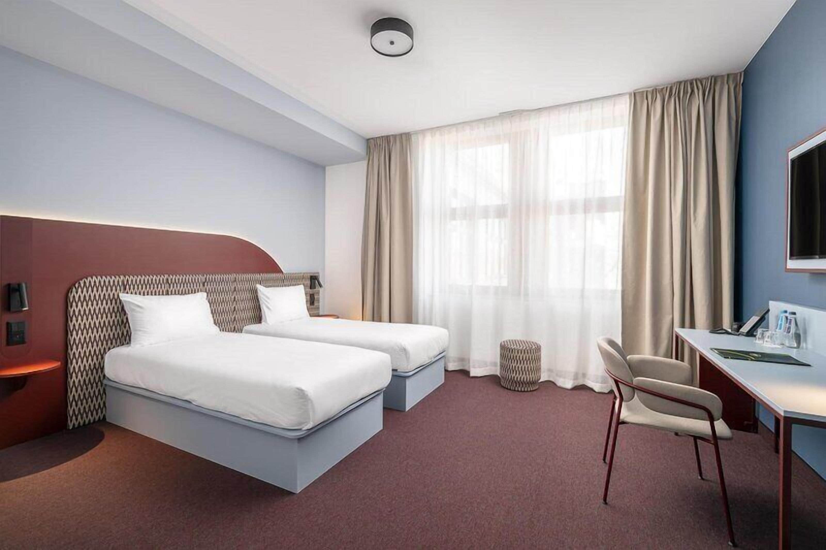 Una camera d'albergo con due letti, una scrivania, una sedia, una TV e una finestra con tende.