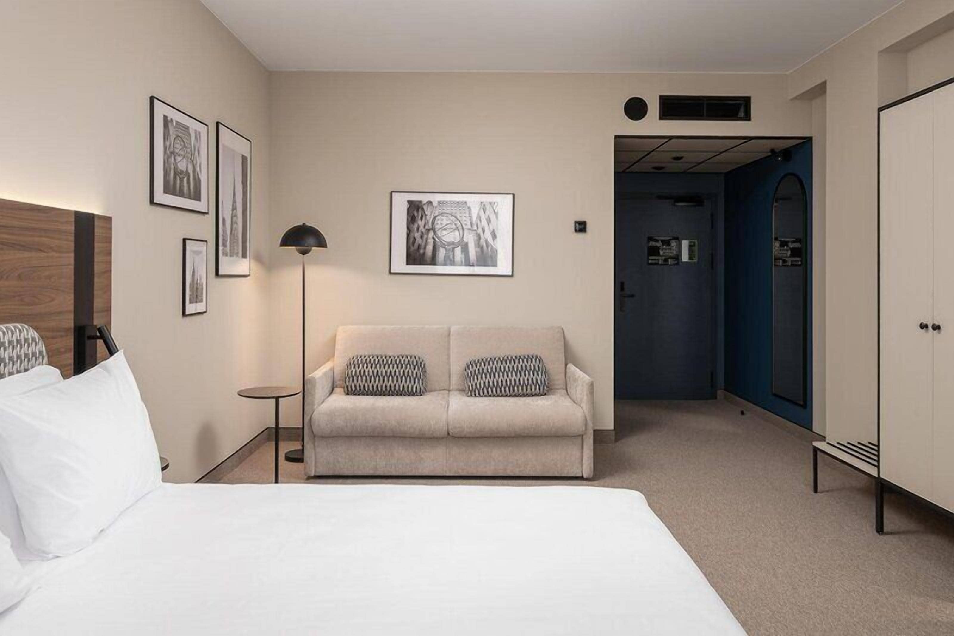 A hotel room with a bed, a sofa, a lamp, framed pictures on the wall, and a wardrobe.