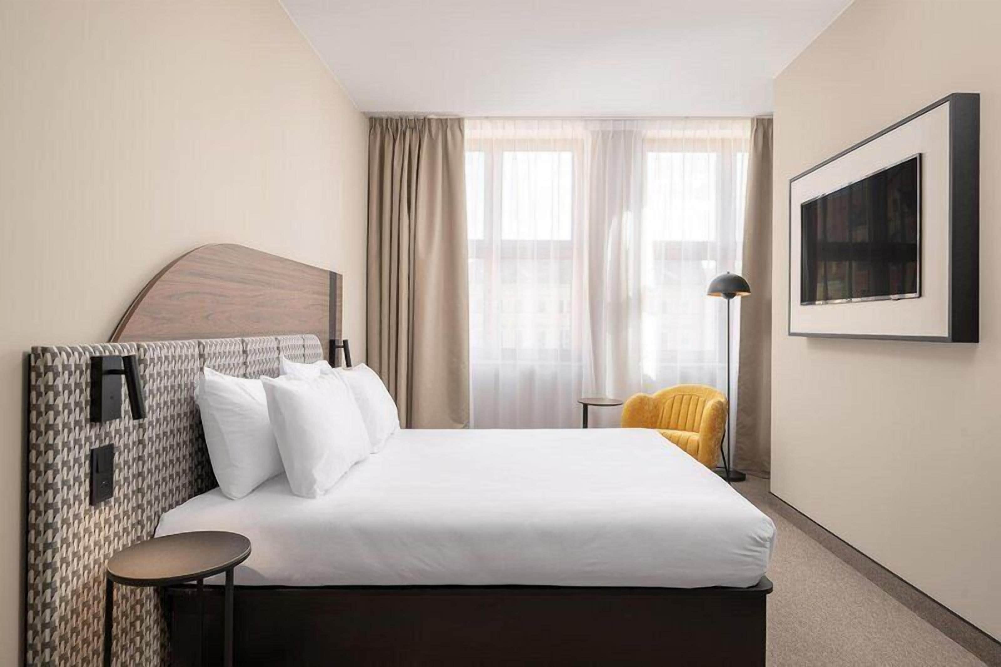 A hotel room with a bed, a round side table, a chair, a floor lamp, and a framed picture on the wall.