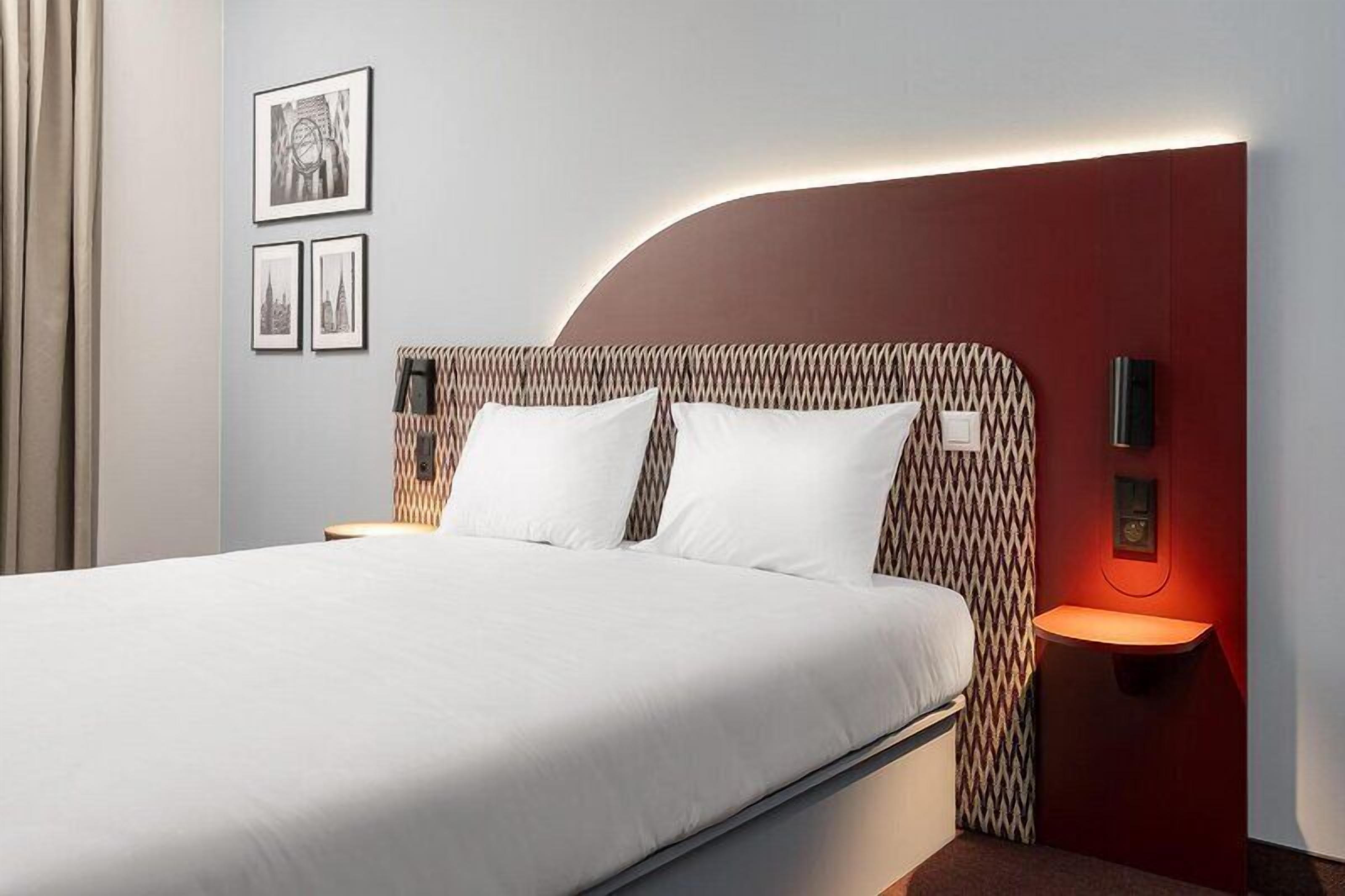 Une chambre d’hôtel avec un lit, deux lampes de chevet, une horloge fixée au mur et des tableaux encadrés accrochés au mur.