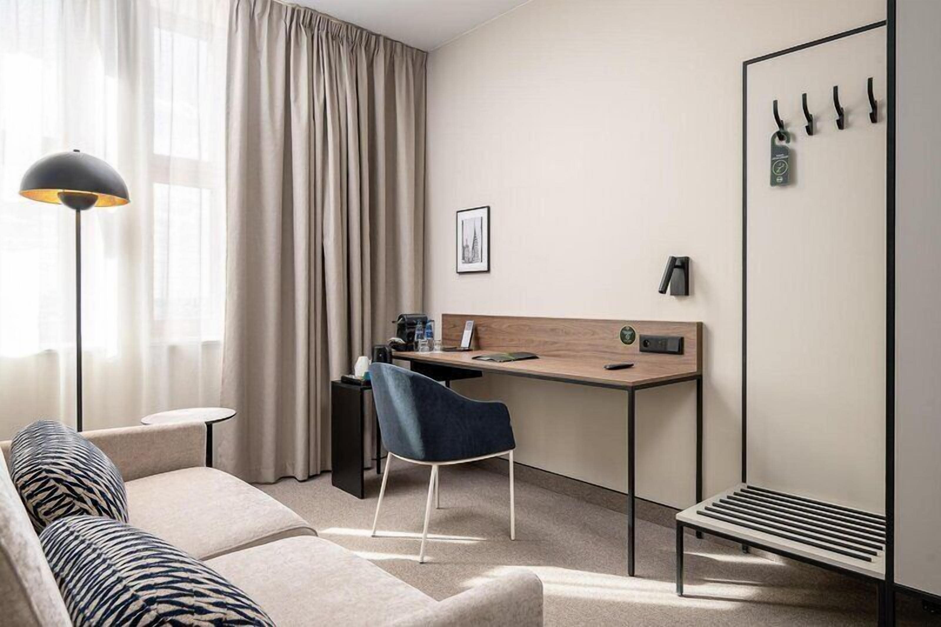 Une chambre d’hôtel moderne équipée d’un canapé, d’un bureau, d’une chaise, d’une lampe, d’un porte-manteau et d’une fenêtre avec des rideaux.