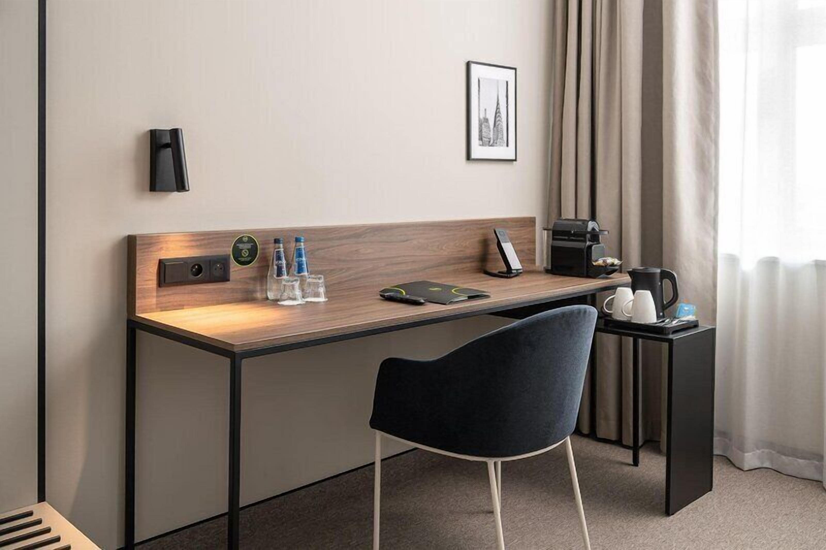 A hotel room with a desk, chair, coffee maker, water bottles, and a framed picture on the wall.