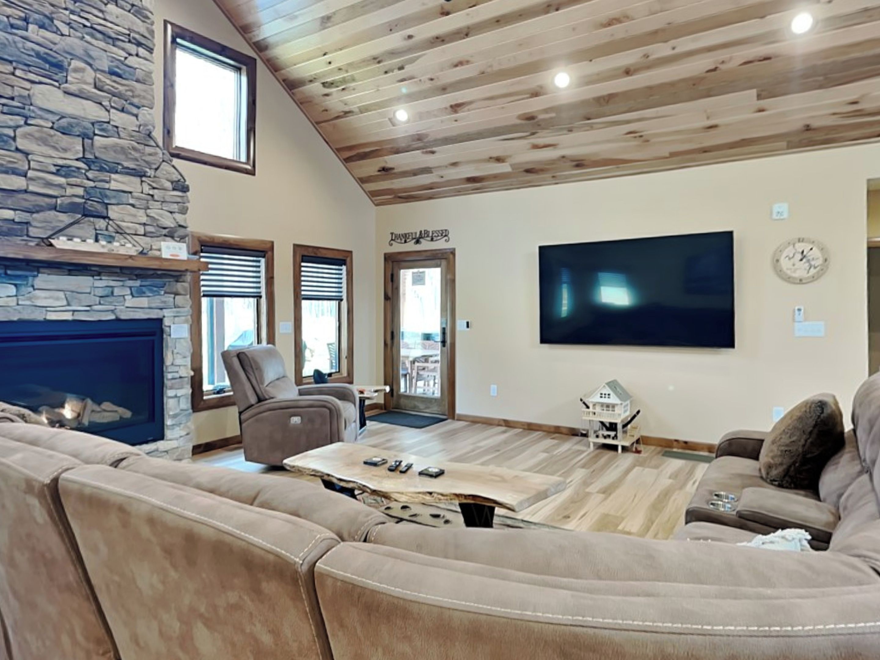 A living room with a stone fireplace, a large flat-screen TV, and a wooden ceiling.
