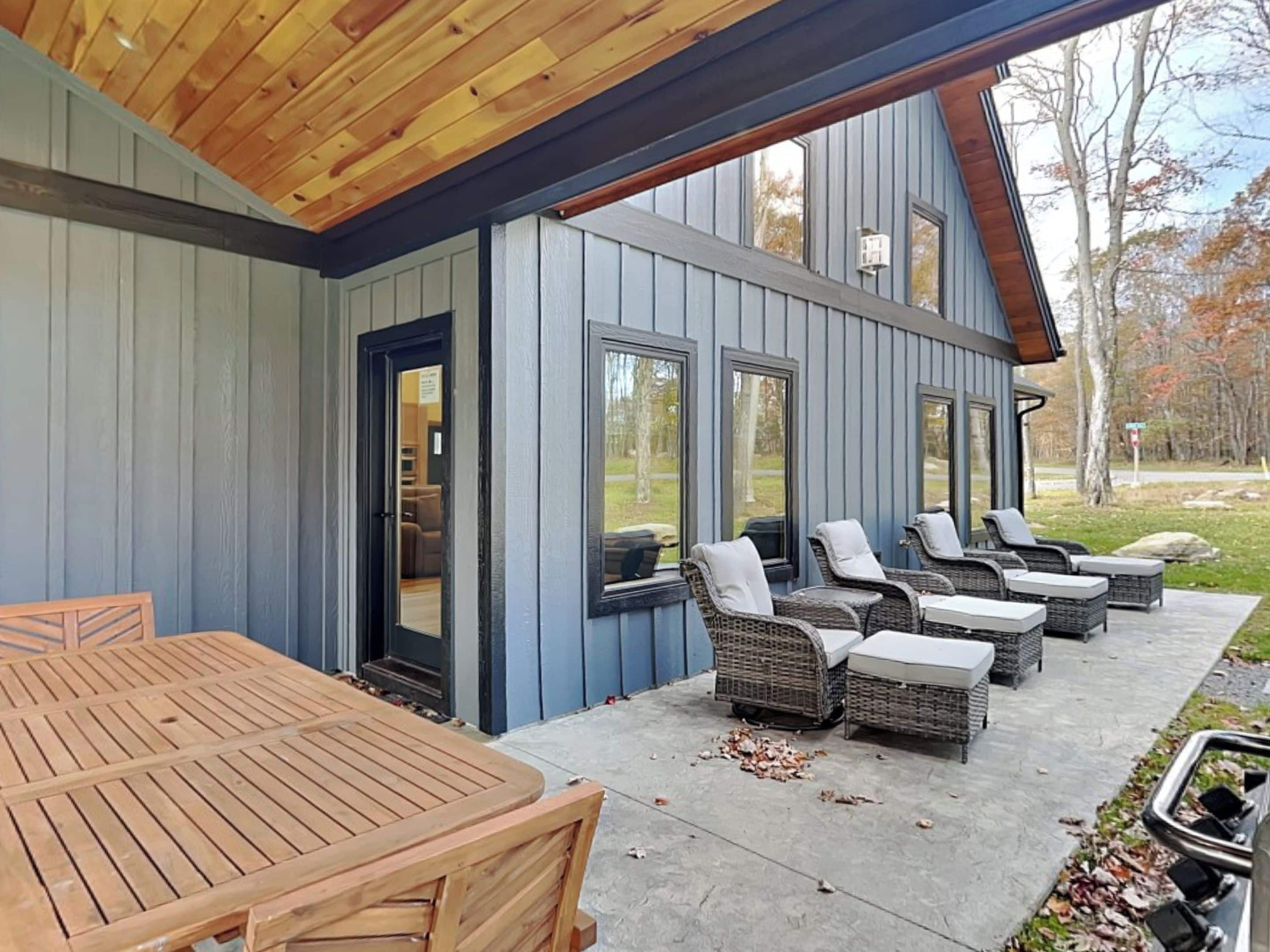 A covered patio with a wooden table and wicker chairs, a blue exterior wall, and large windows.