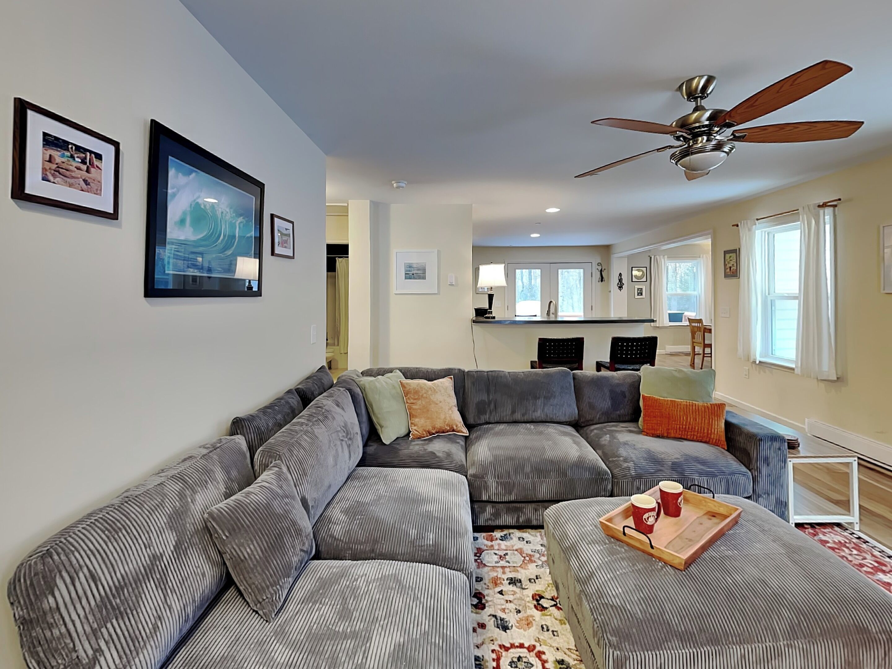 A living room with a sectional sofa, a ceiling fan, and a kitchen area in the background.