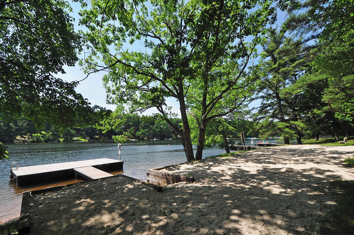 A lakeside park with a wooden dock extending into the water, surrounded by trees and a sandy area with benches.