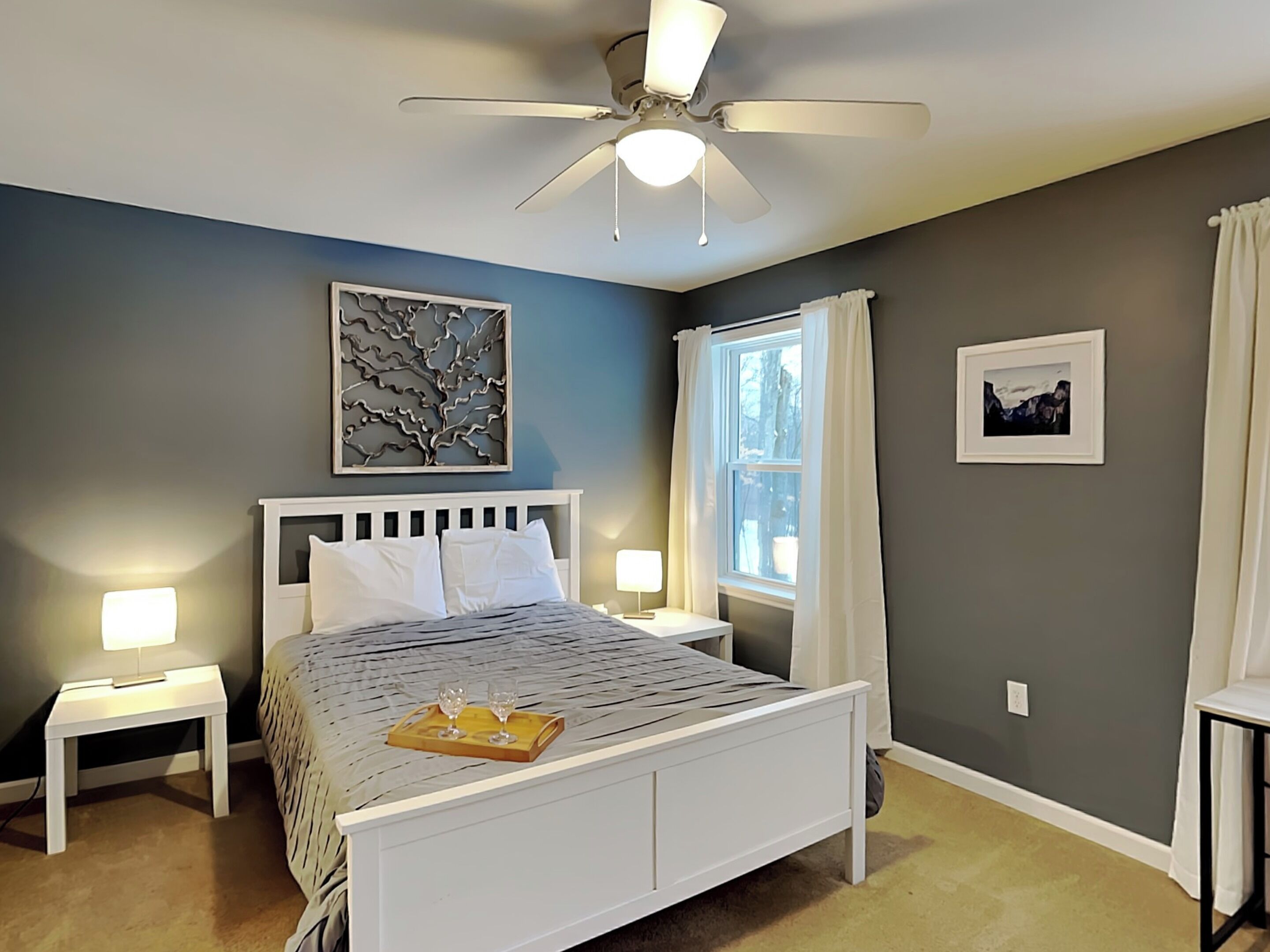 A bedroom with a bed, bedside tables, a ceiling fan, a window with curtains, and wall art.