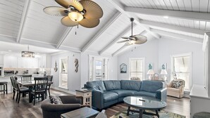 A spacious living area with a large sectional sofa, a glass coffee table, and a dining table with chairs. There are ceiling fans and pendant lights.