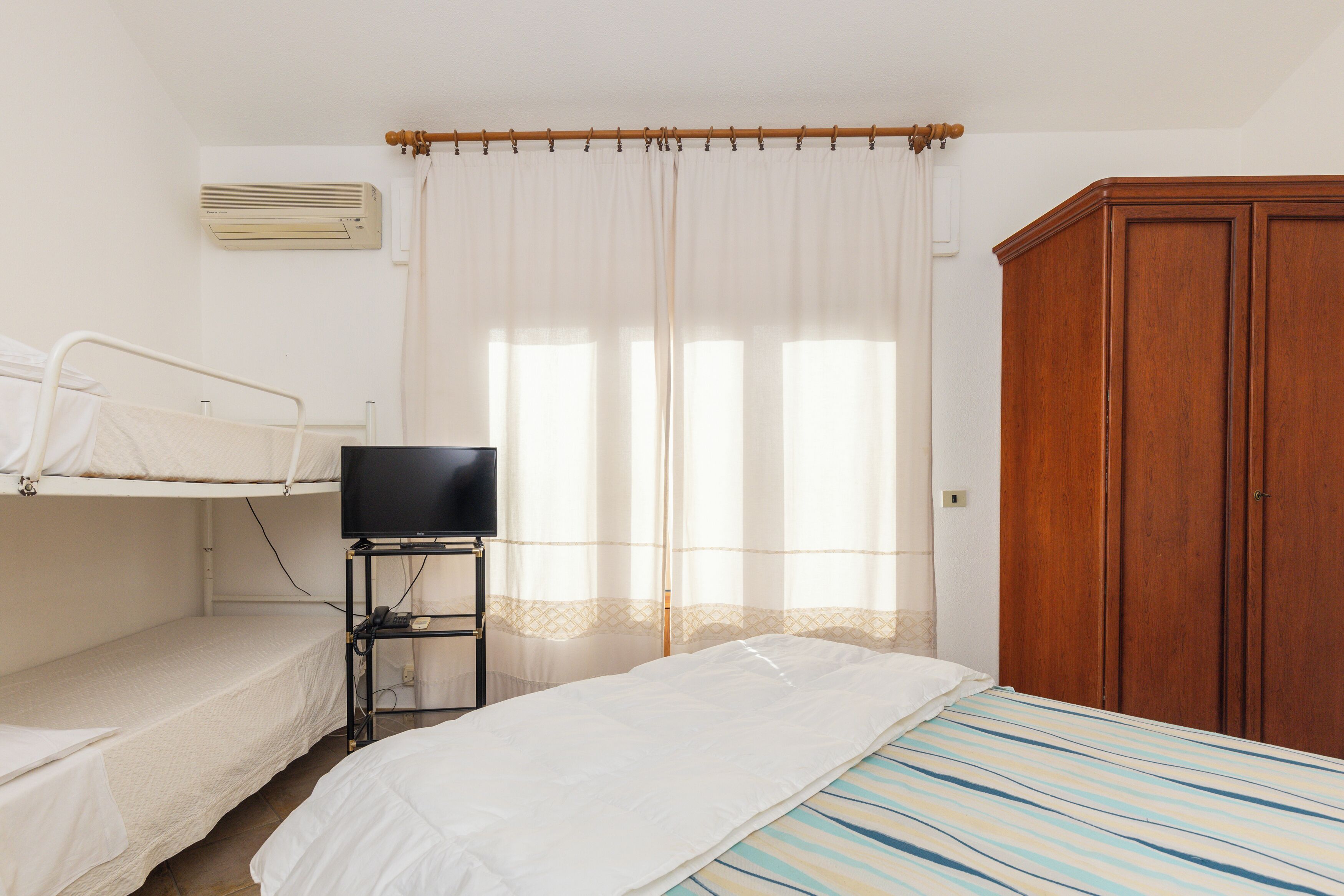 A room with bunk beds, a television on a stand, a wooden wardrobe, and a window with sheer curtains.
