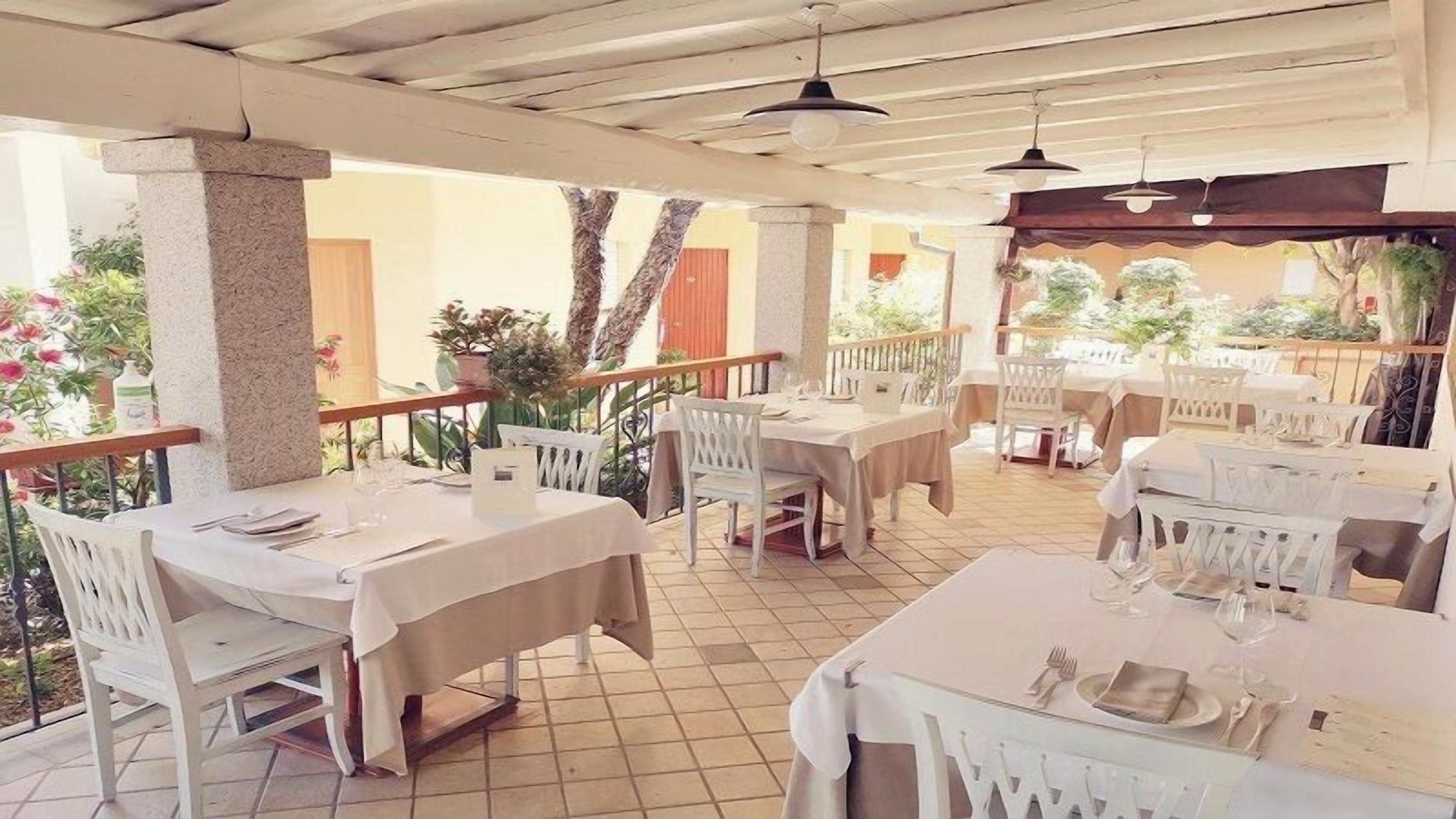 Un restaurante al aire libre con mesas y sillas blancas, piso de baldosas y una zona cubierta con baranda de madera.