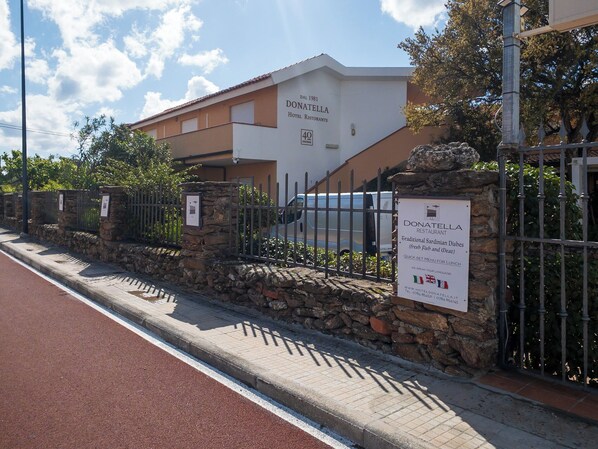A hotel entrance with a stone wall, metal gate, and a signboard advertising traditional Sardinian dishes.