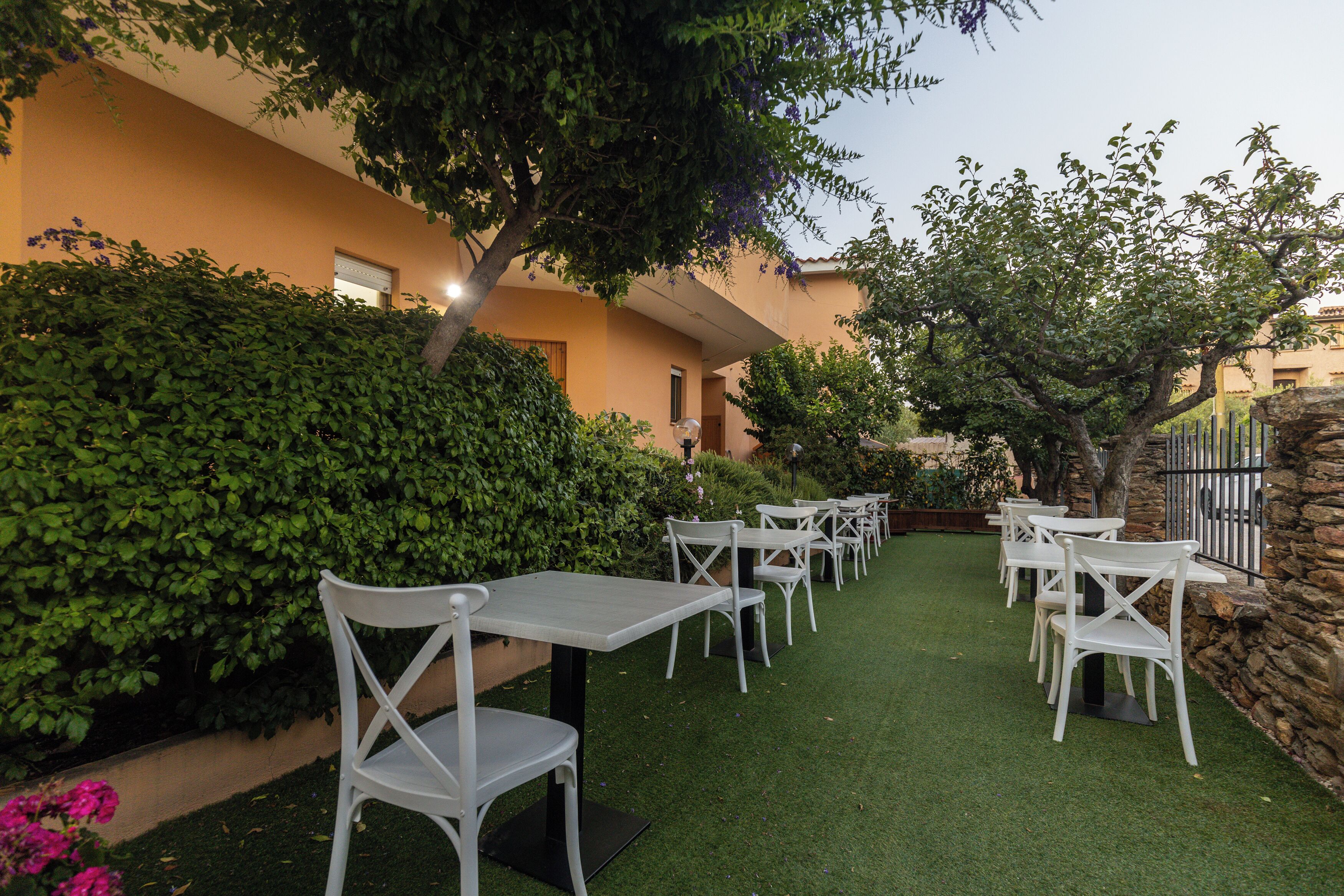 An outdoor seating area with white tables and chairs, surrounded by greenery and a stone wall.