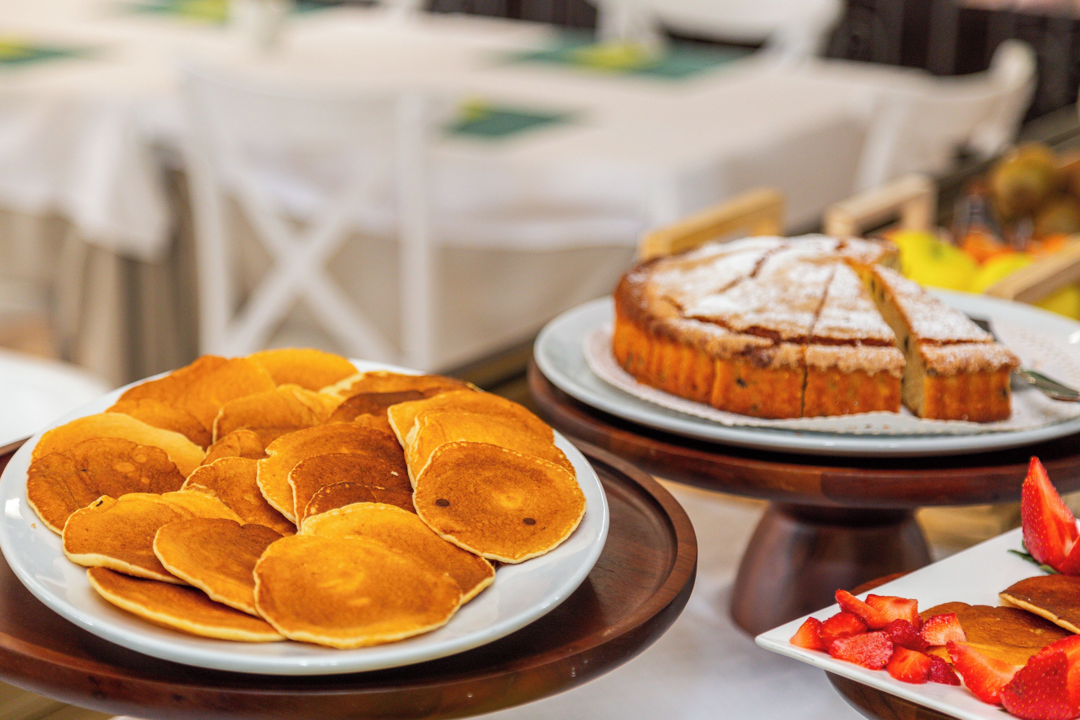 Uma mesa com pratos de panquecas e um bolo, com uma travessa de frutas em primeiro plano.