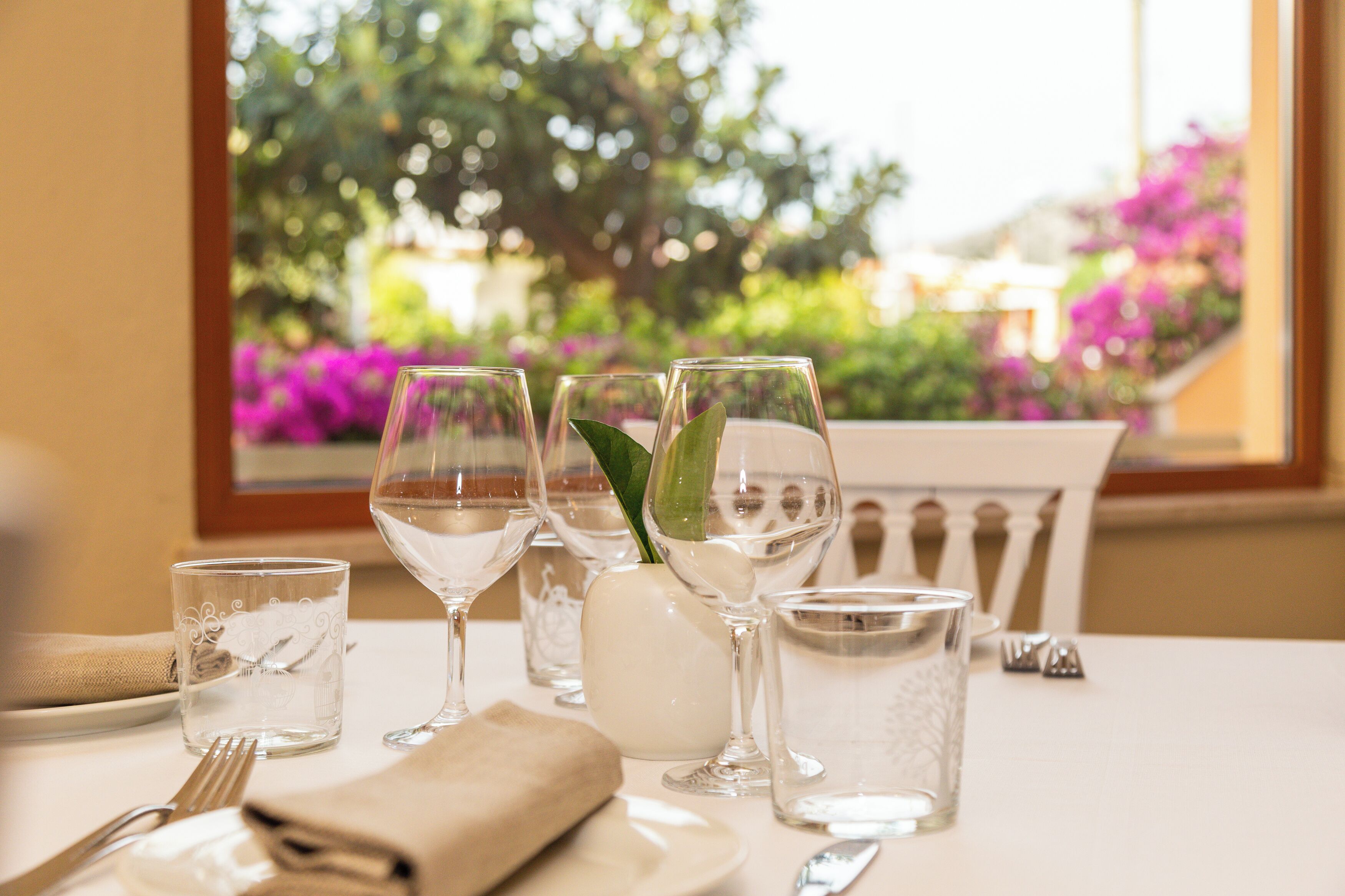 A dining table set with clear glassware, a white vase with a green plant, and a view of a garden with purple flowers through a window.