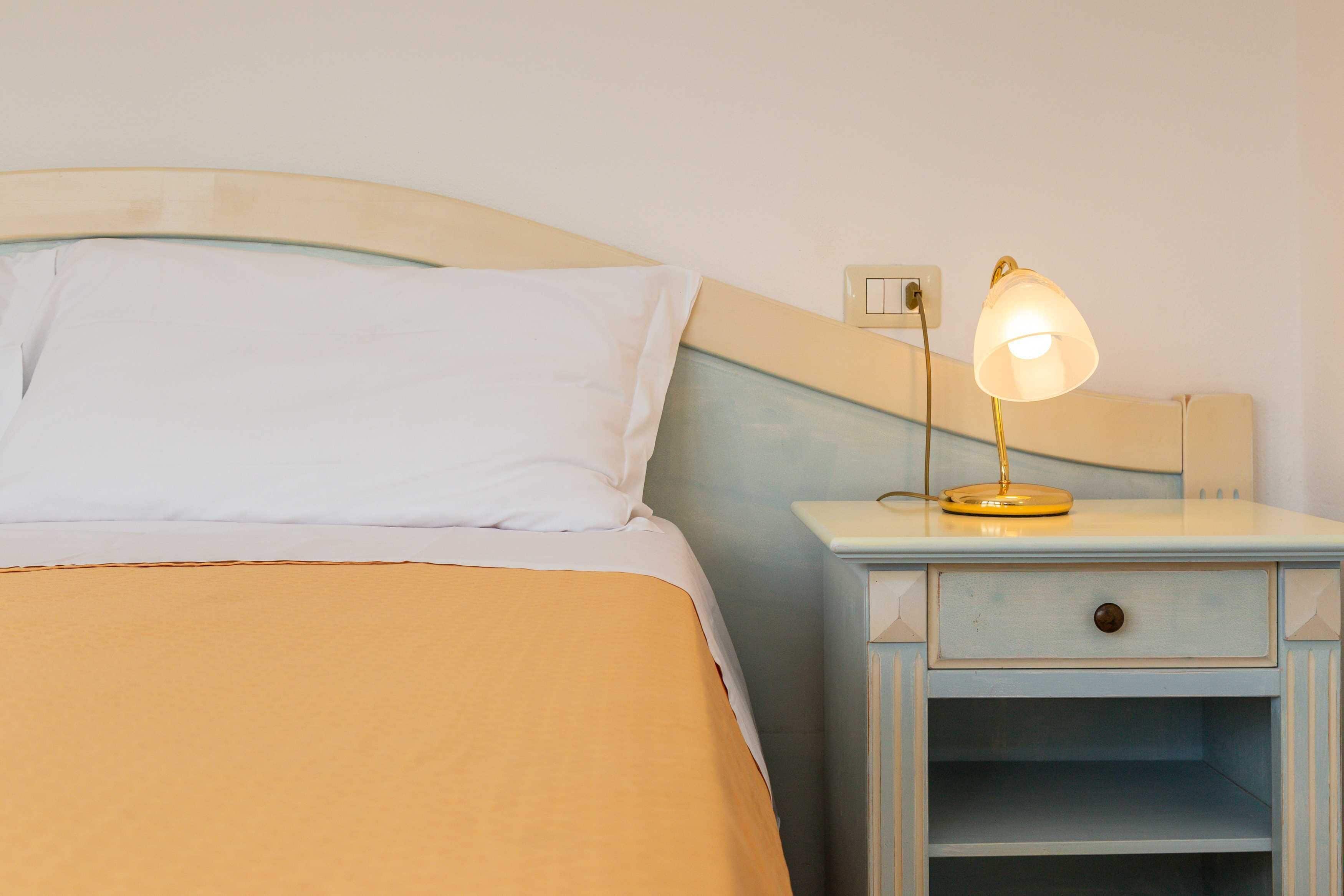 Ein ordentlich bezogenes Bett mit einer weiß-beigen Bettdecke, ein Nachttisch mit Lampe und eine Schublade.
