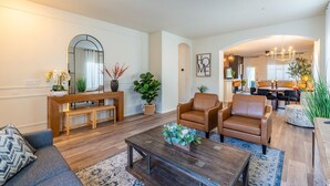 Un salon moderne avec un canapé, une table basse et deux fauteuils. On y trouve également un coin repas avec une table et des chaises, et une cuisine à l’arrière-plan.