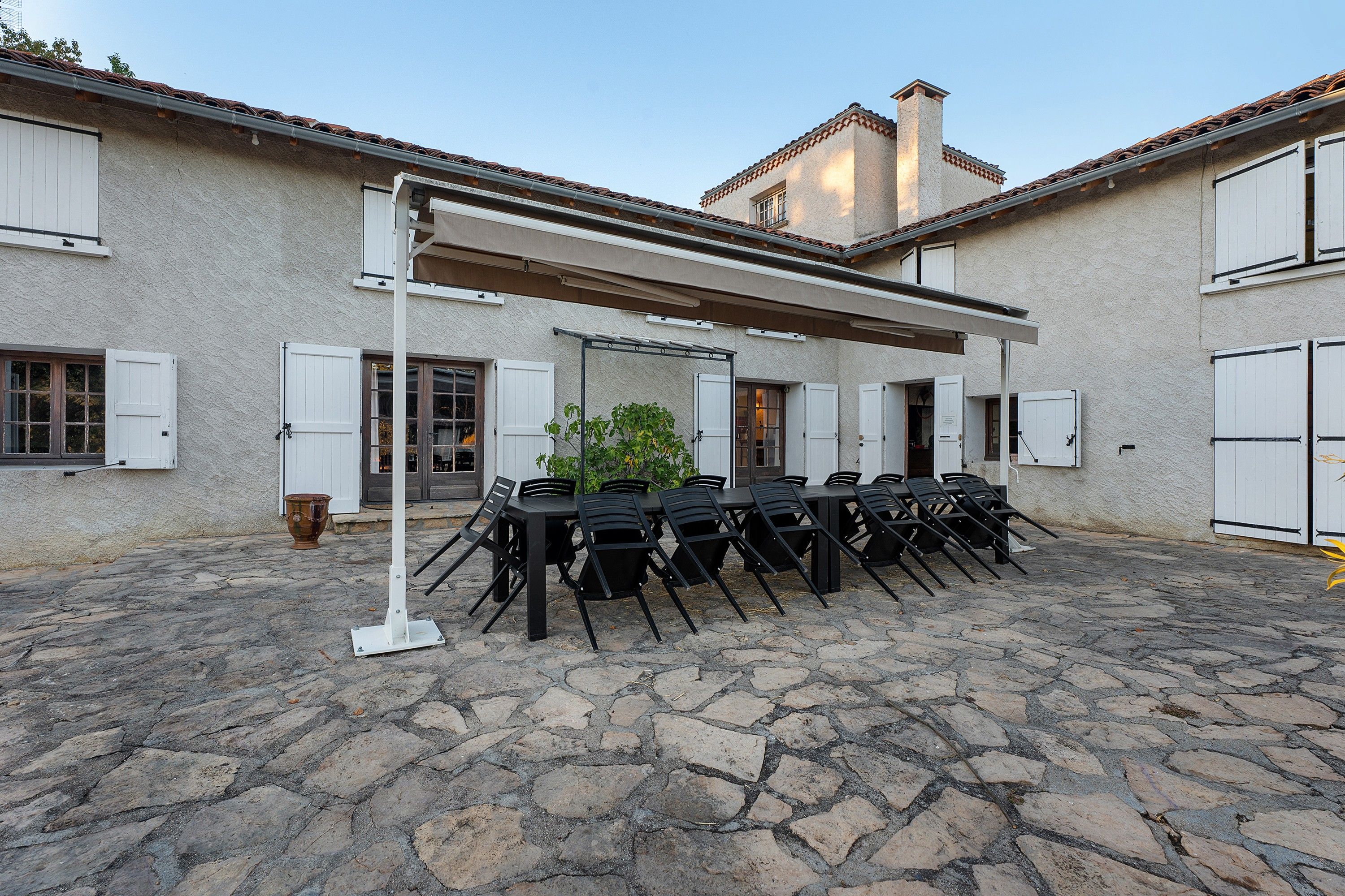 An outdoor patio with stone flooring, a covered seating area with tables and chairs, and a building with multiple windows and doors.