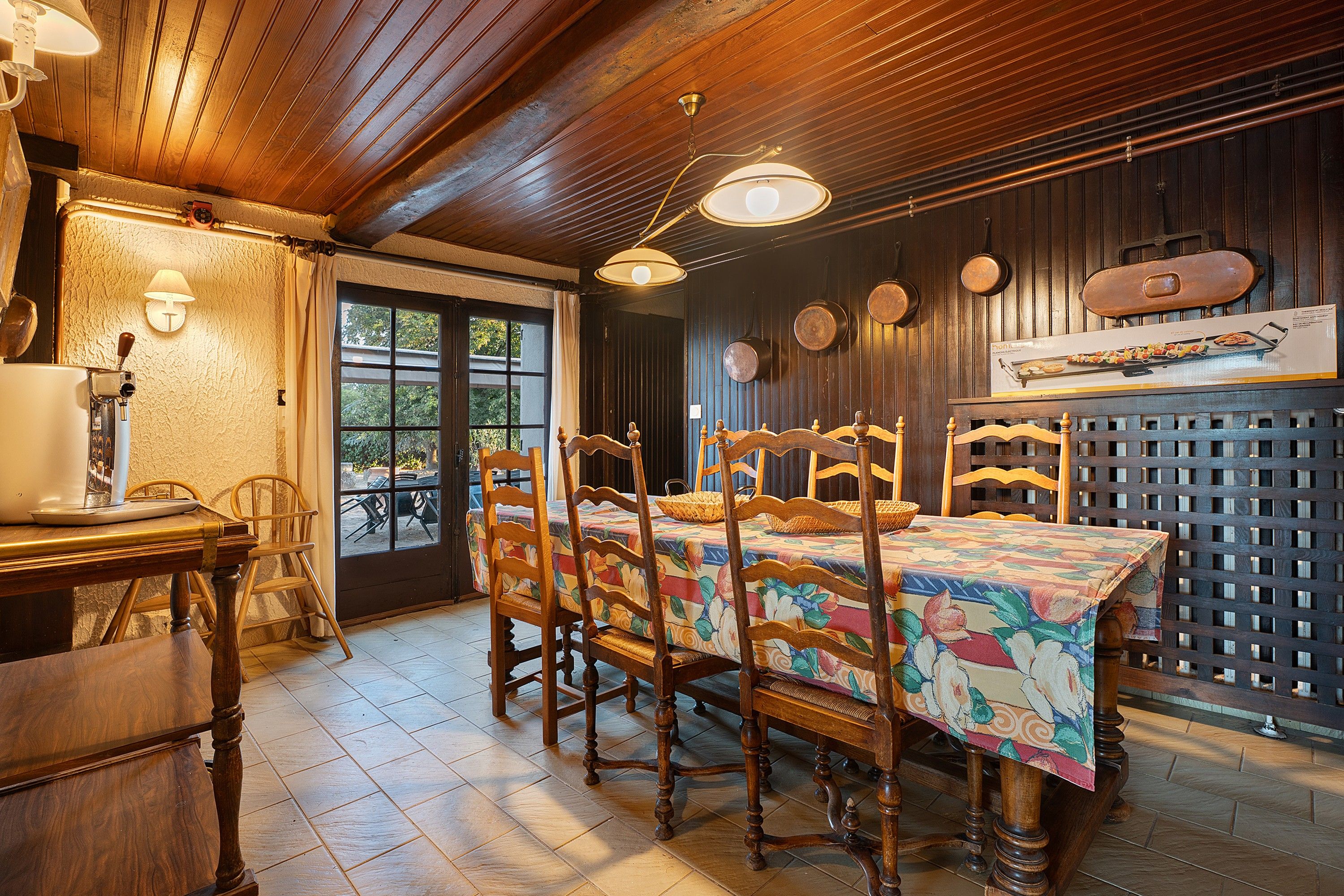 Un coin repas avec des tables et des chaises en bois, une fenêtre avec des rideaux et un porte-ustensiles fixé au mur.