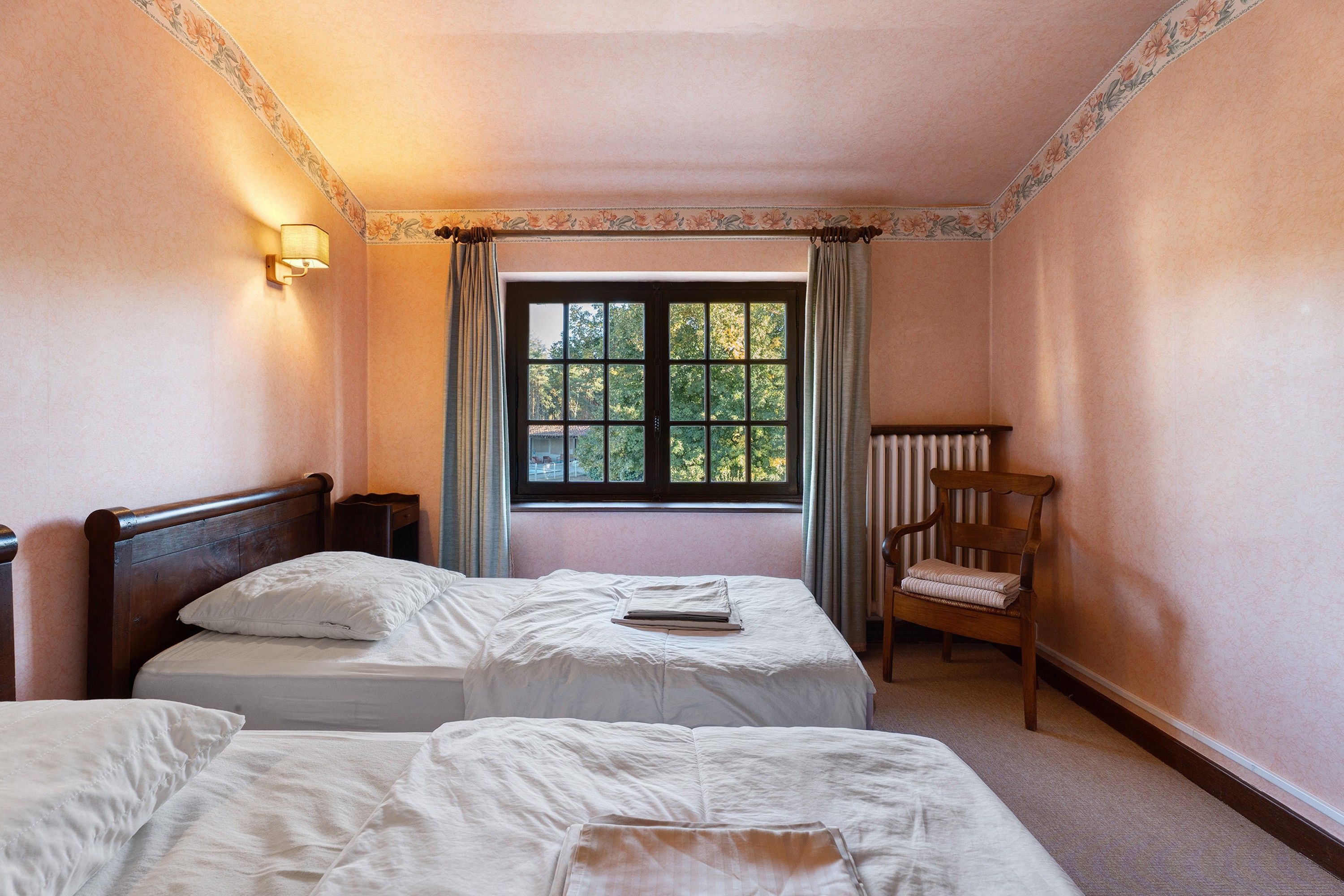 A hotel room with two beds, a chair, a window with a view of trees, and a radiator.