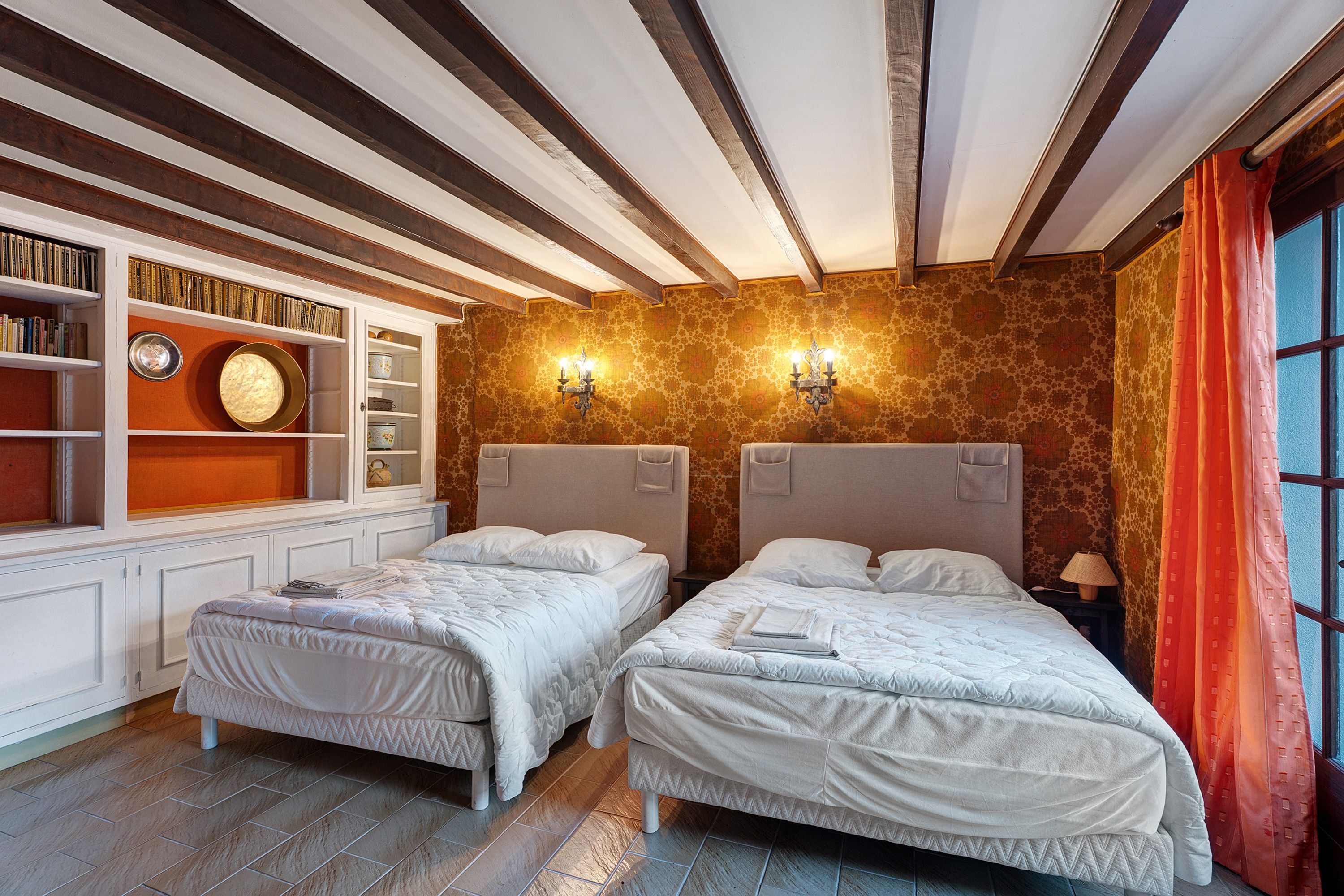 A room with two beds, a bookshelf, a clock, and a window with curtains.