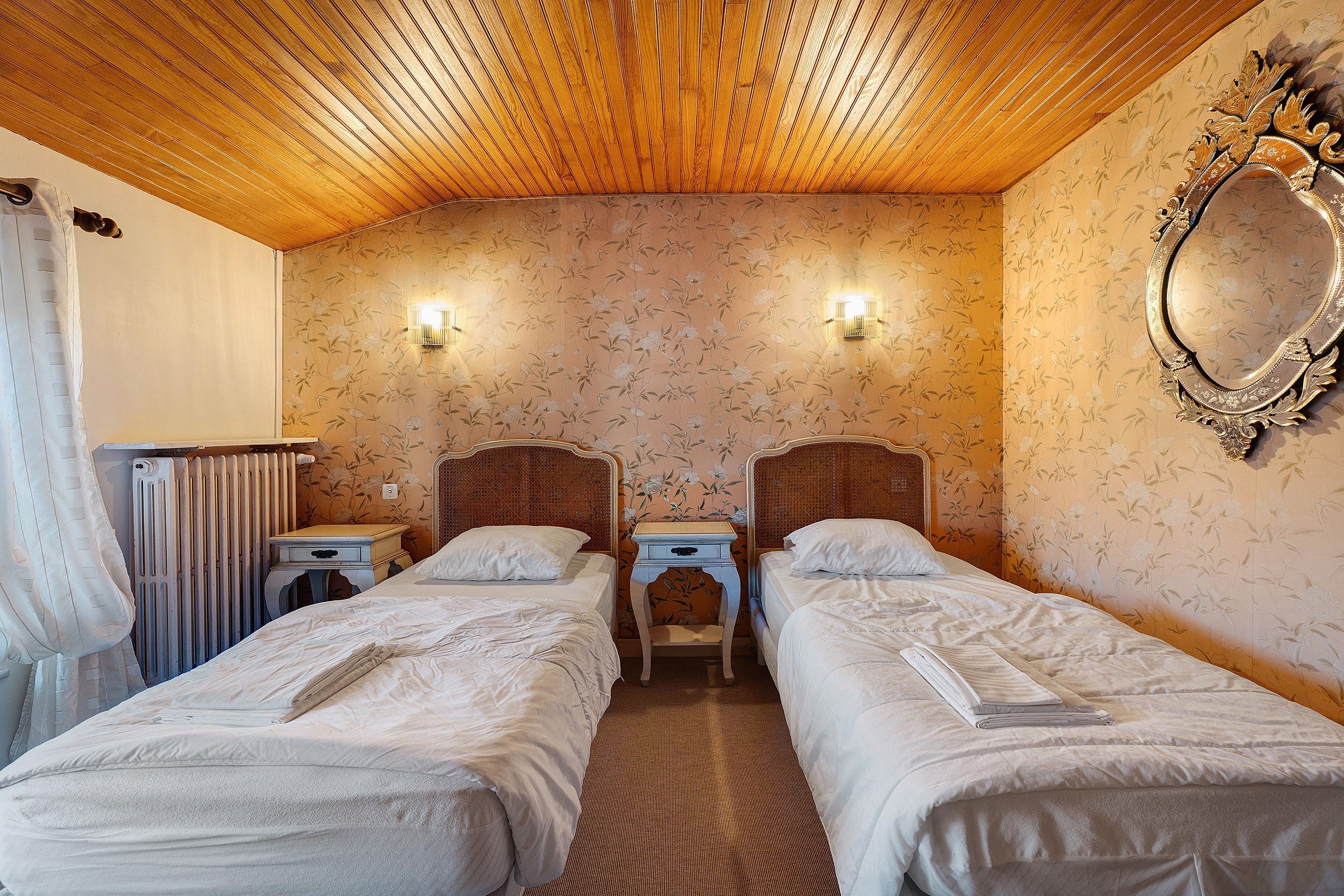 Habitación de hotel con dos camas individuales, techo de madera, papel tapiz floral, un espejo y un radiador.
