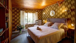 A bedroom with a wooden ceiling, patterned wallpaper, a bed with pillows, a bedside table with a lamp, and a window with curtains. - Gîte des roties: Restored Family Home with Pool in a 7-Hectare Park (Poncins)