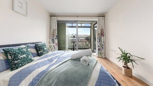 A bedroom with a bed, pillows, a rolled towel, a wooden bedside table with a plant, and a view through a glass door. - *Modern Spacious 1 Bedroom Flat in Camden* (London)