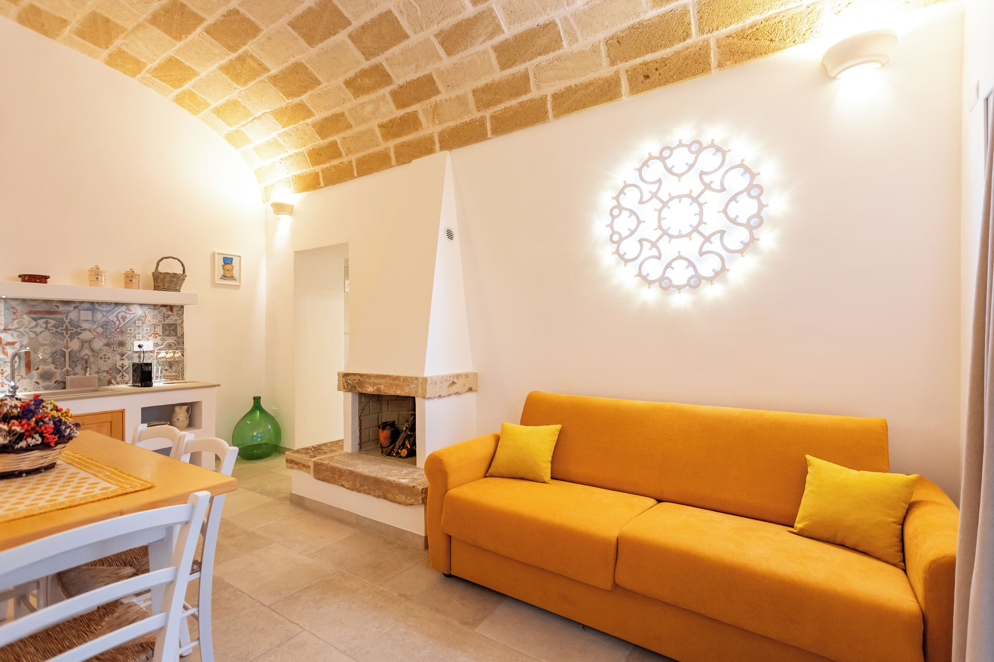 A living room with a yellow sofa, a fireplace, and a kitchen area with a tiled backsplash.