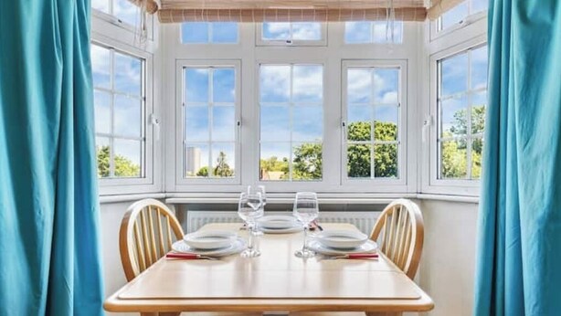 A dining area with a table set for four, large windows with curtains, and a view of greenery outside. - *Modern Spacious studio flat in Ealing* (Hangerlane )