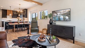 A modern living room with a large wooden dining table, a leather sofa, and a cowhide rug. There is a black sofa, a coffee table with a vase, and a painting of a lake on the wall. - Walk to Town | Near Free Shuttle (Fraser)