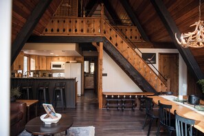 A spacious loft-style living area with a high ceiling, wooden beams, a central staircase, and a kitchen in the background. - 🔥 Cozy Bellaire A-Frame | Golf • Torch • Brew 🍻<br>Nestled in Schuss Mountain Resort—close to Golf, Torch Lake 💙, and downtown Bellaire’s famous Short’s Brewery. Stay, play, and unwind by the fire pit under Northern Michigan skies 🌲✨ (Bellaire)