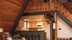 Un espacio habitable amplio con techo alto y vigas de madera, un rincón de descanso acogedor con un sofá de cuero y una mesa de centro, y una cocina al fondo.
