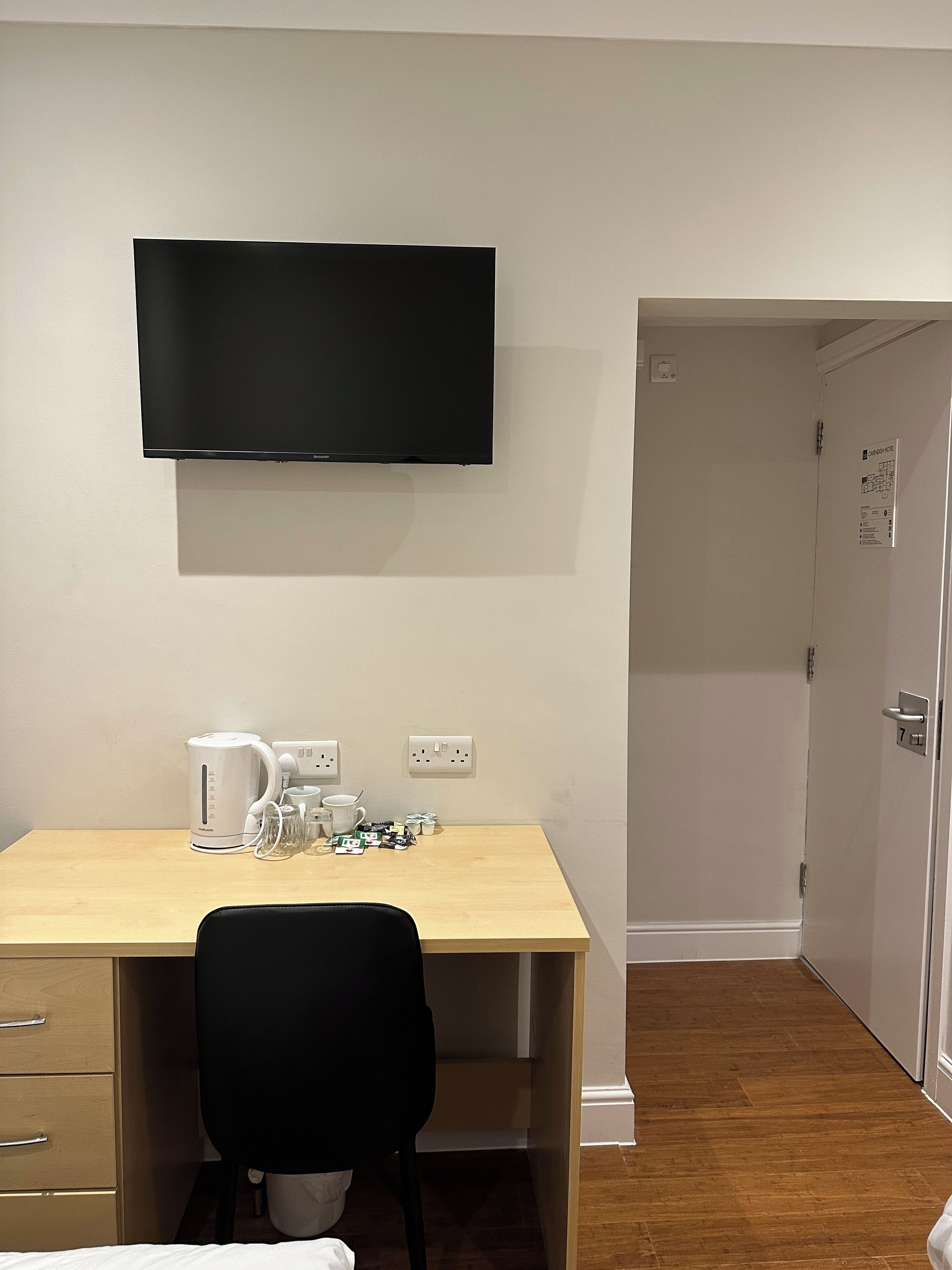 A room with a wooden desk, a black chair, a flat-screen TV mounted on the wall, a kettle, and a coffee maker.