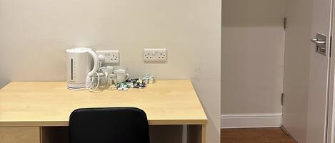 A room with a wooden desk, a black chair, a flat-screen TV mounted on the wall, a kettle, and a coffee maker.